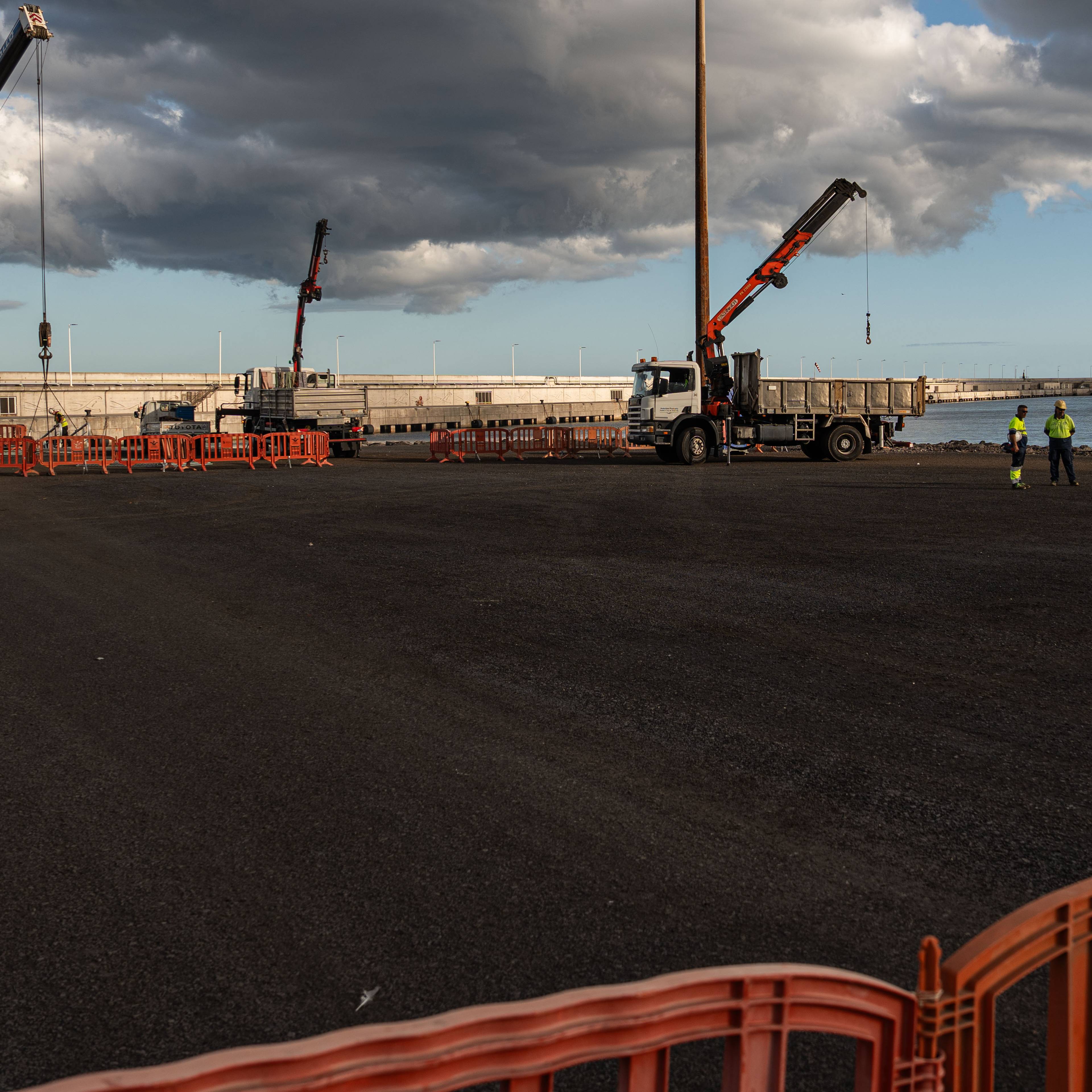 Arbeidere i gul og grønn vest står ved en havn. Flere kraner og lastebiler er synlige på anleggsområdet. Et stort skip ligger til kai i bakgrunnen. (Bildebeskrivelsen er laget av en KI-tjeneste)
