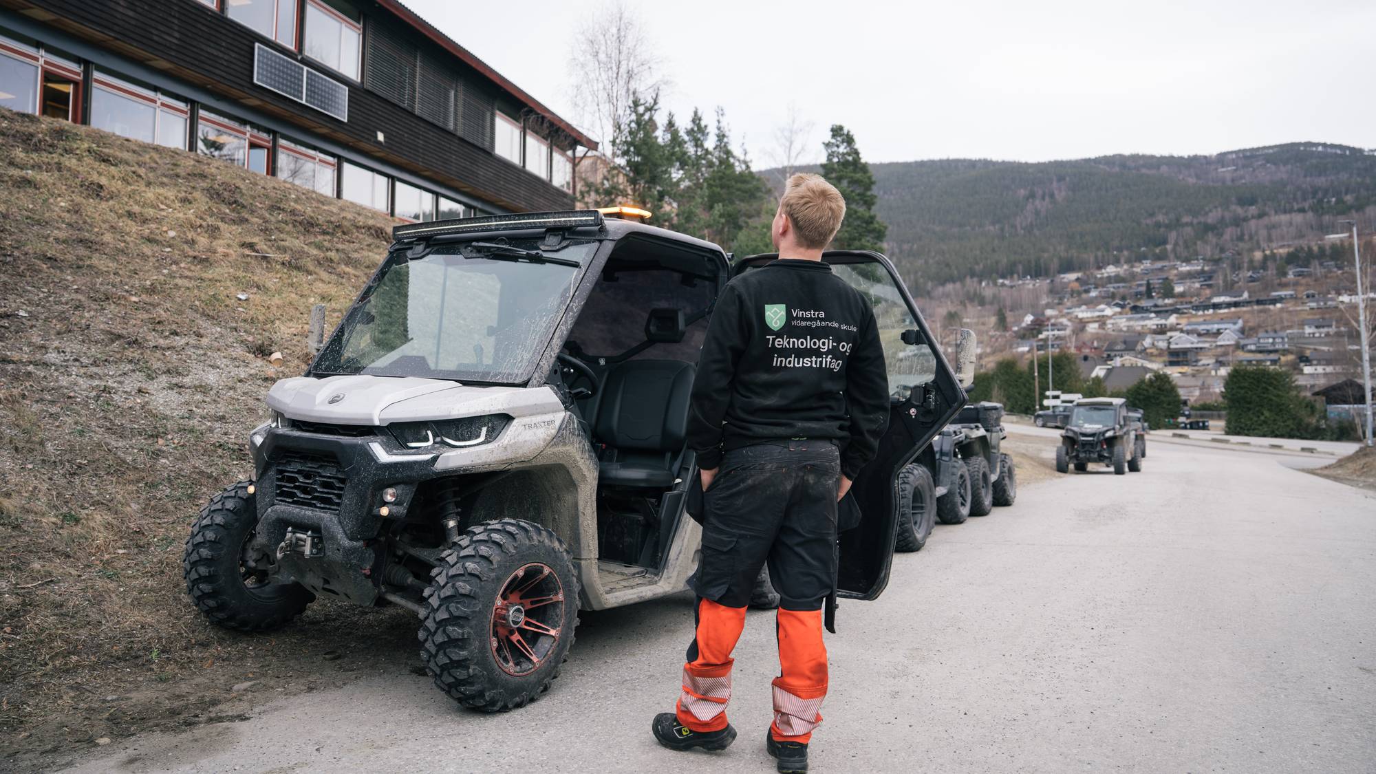 En ung mann står ved siden av en firehjuling. Han har på seg en genser med logoen til Vinstra videregående skole. Bak ham står det flere UTV-er og firehjulinger på en vei. 