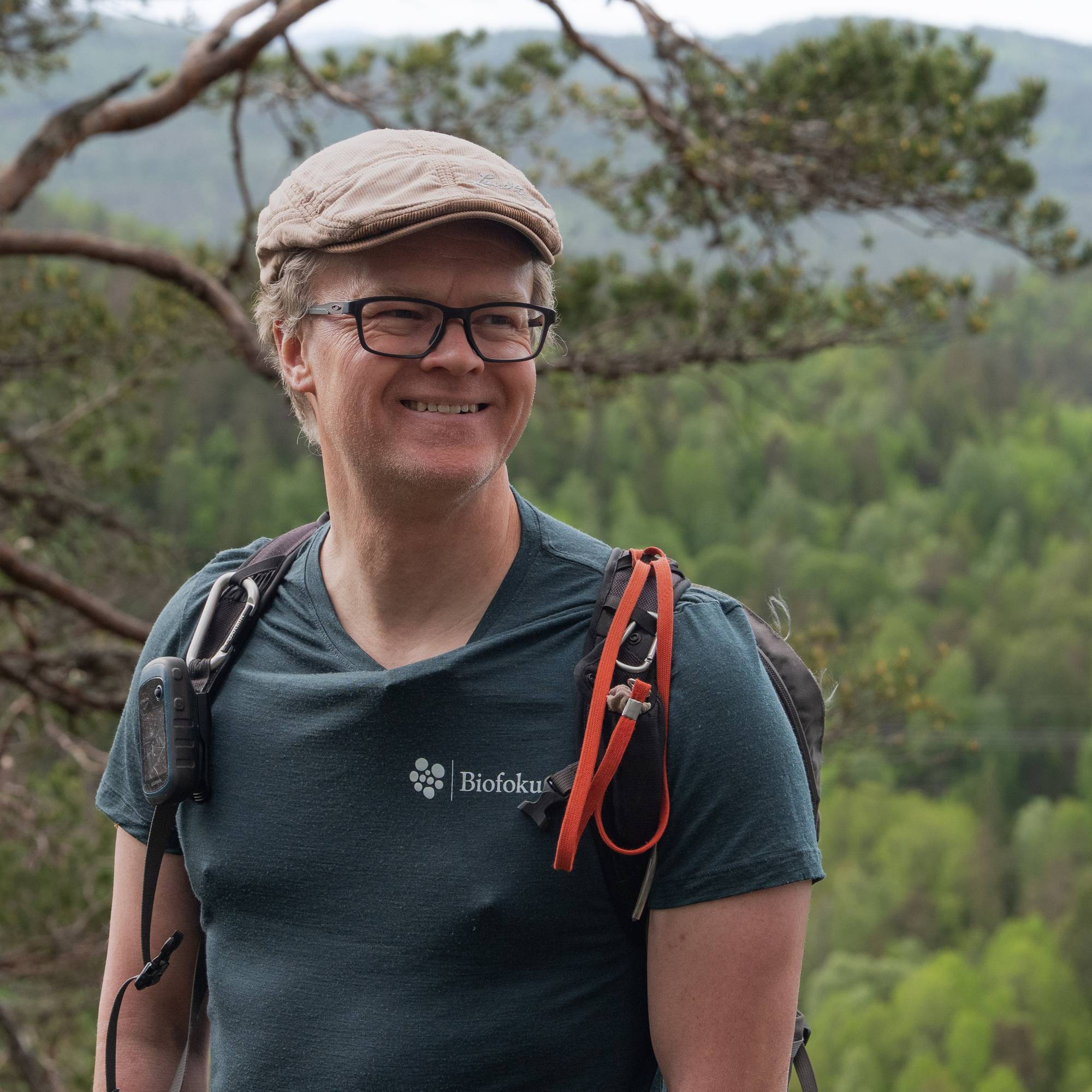 Mann med sixpence, briller og t-skjorte med skog i bakgrunnen. 