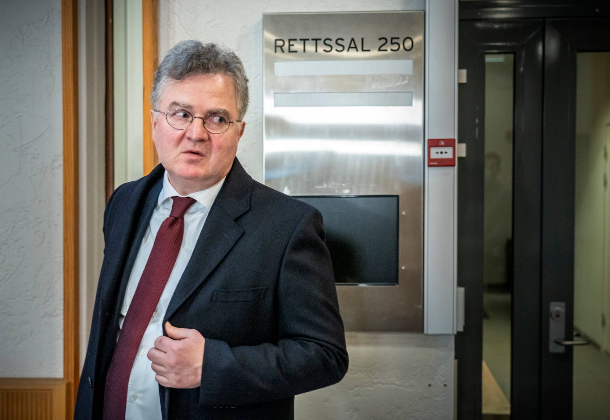 En mann i dress og slips står foran en dør med skiltet RETTSAL 250. Mannen har briller og ser mot høyre. Han holder en hånd på jakkekragen. En brannalarm er festet til veggen ved siden av skiltet. (Bildebeskrivelsen er laget av en KI-tjeneste)