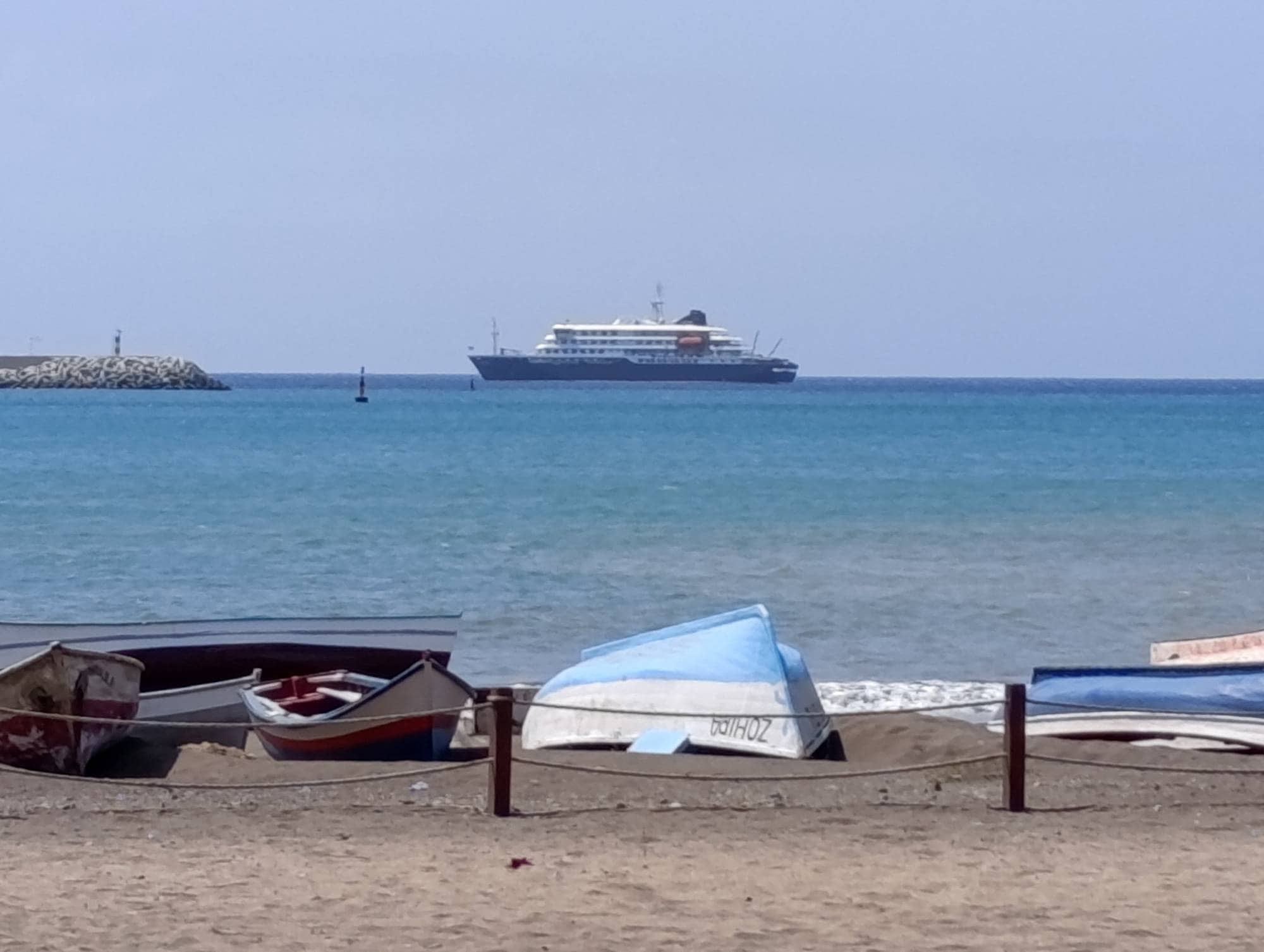 MV Hondius - Sandstrand med en trebåt som ligger med blå bunn i været, ute på sjøen ligger et lite cruiseskip