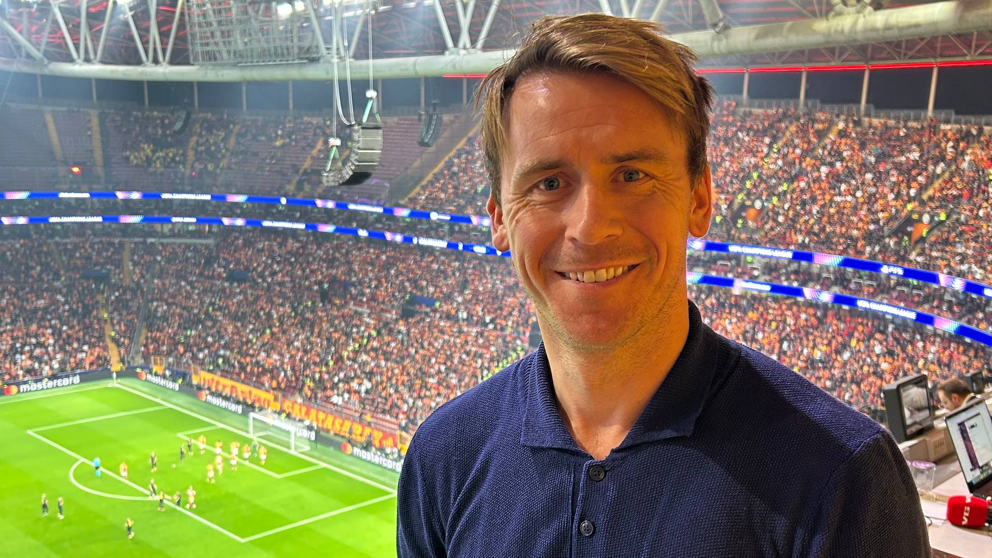 En person står foran en sportsarena med stor publikum. I bakgrunnen er et fotballspill i gang på banen. Det er mange tilskuere samlet, og stemningen er intens. Området er opplyst av stadionlys.