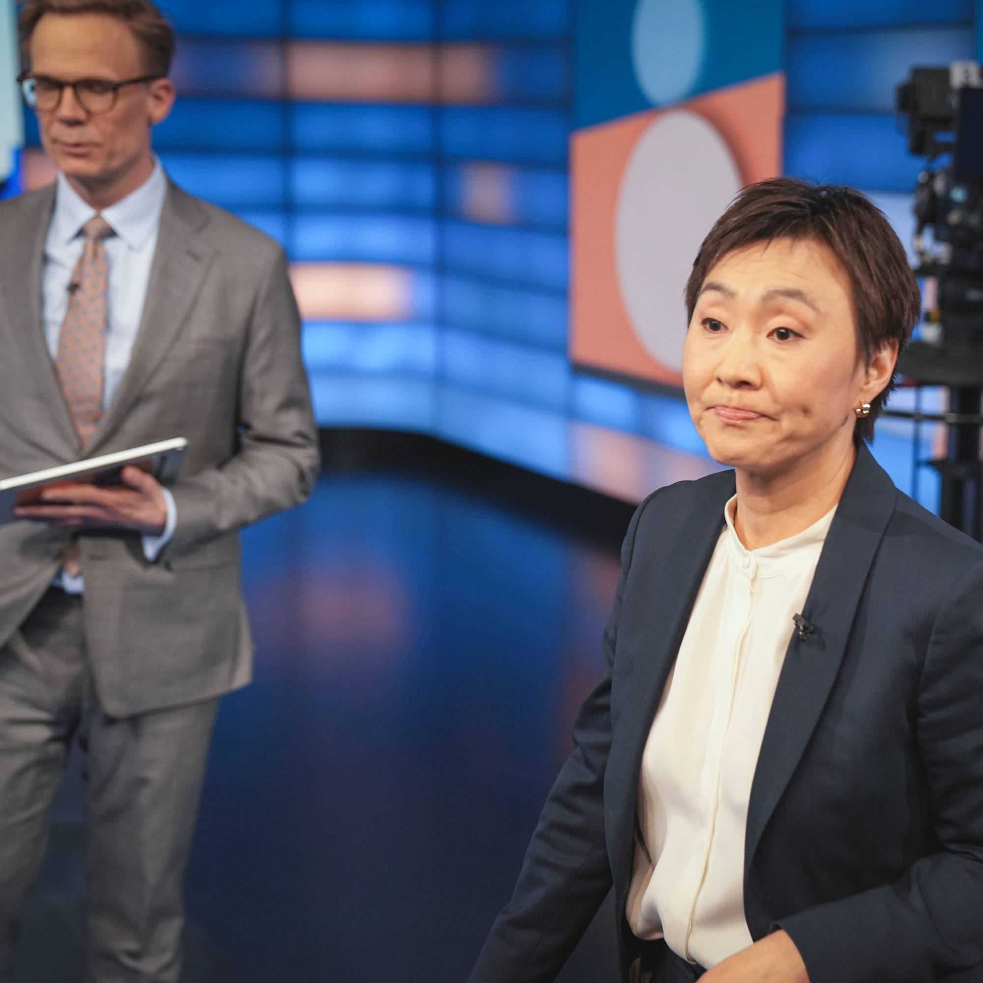 En kvinne i mørk dress står i forgrunnen, mens en mann i lys dress står i bakgrunneni studio til NRK Debatten