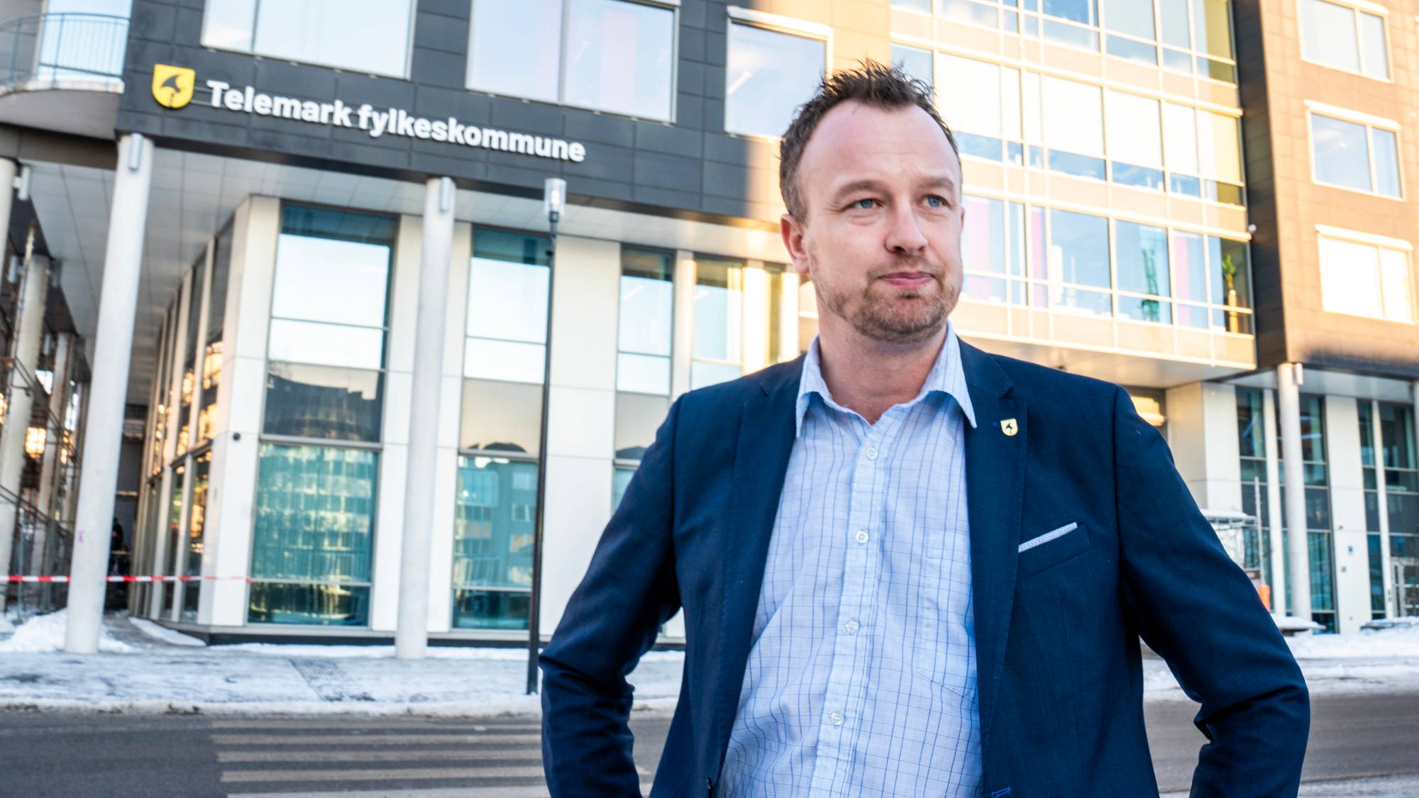 en mann med dressjakke og skjorte som holder henda på hoftene foran et stor bygg der det står Telemark fylkeskommune på forsiden.