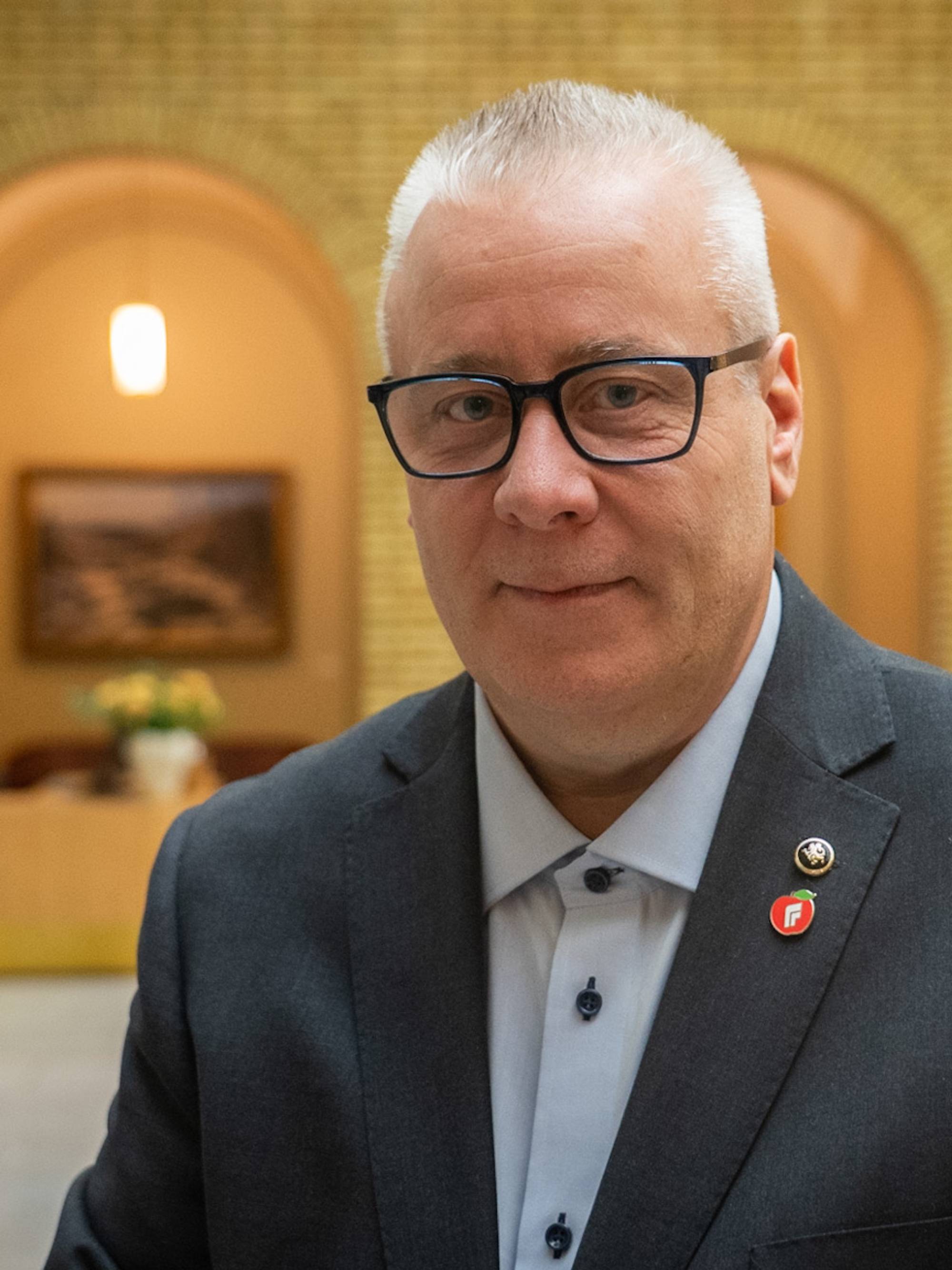 En mann i en mørk dress står i en korridor med mursteinsvegger og kunstverk på veggene. Bakgrunnen viser et mottaksområde med møbler. Mannen har på seg en skjorte med knapper og bærer flere insignier på jakkeslaget. Belysningen i rommet er varm og innbydende. (Bildebeskrivelsen er laget av en KI-tjeneste)