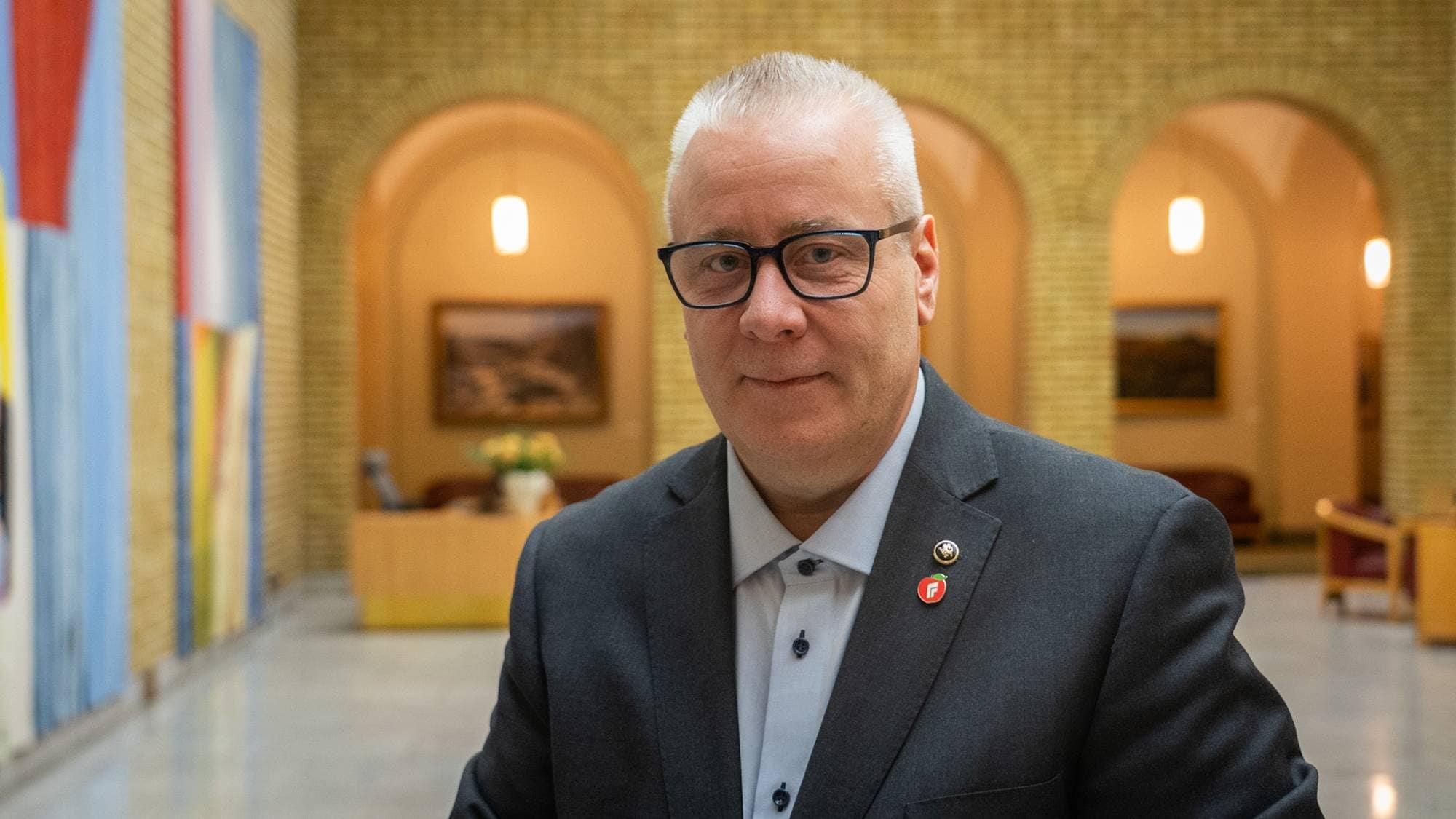En mann i en mørk dress står i en korridor med mursteinsvegger og kunstverk på veggene. Bakgrunnen viser et mottaksområde med møbler. Mannen har på seg en skjorte med knapper og bærer flere insignier på jakkeslaget. Belysningen i rommet er varm og innbydende. (Bildebeskrivelsen er laget av en KI-tjeneste)