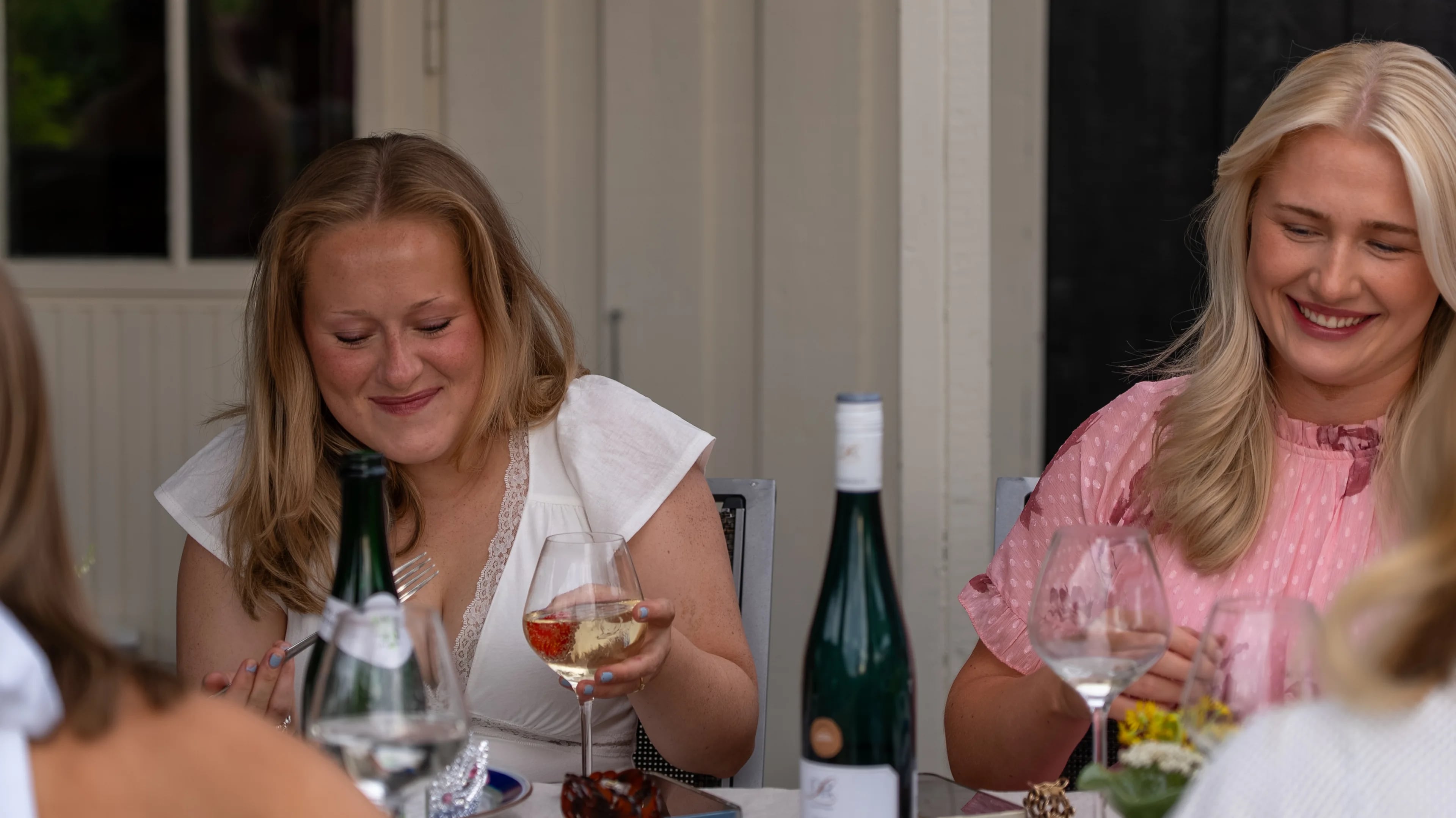 To kvinner sitter ved et bord med glass og vinflasker utendørs. (Bildebeskrivelsen er laget av en KI-tjeneste)