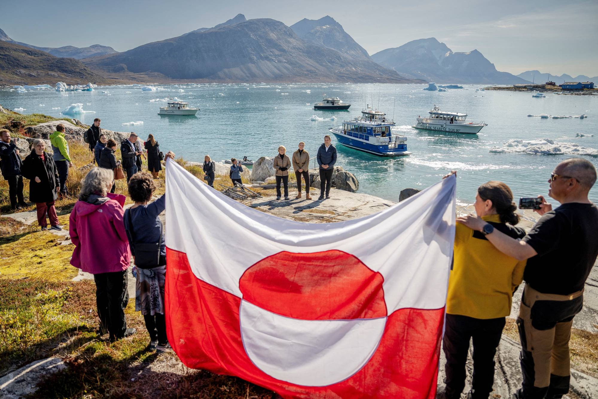 Folk holder Grønlands flag foran Ursula von der Leyen og Mette Frederiksen ved en fjord i Grønland. (Bildebeskrivelsen er laget av en KI-tjeneste)