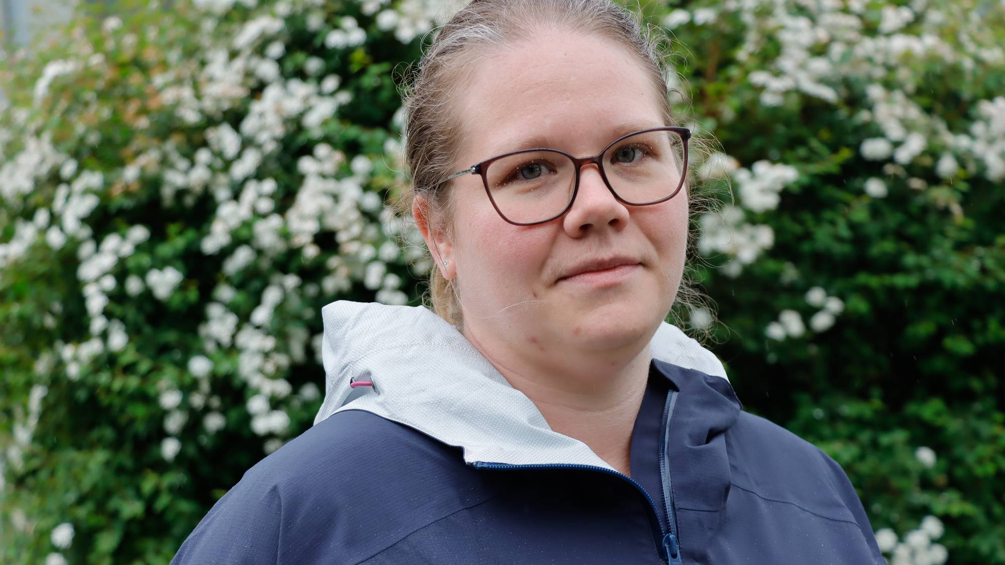 Et bilde av en kvinne med briller og mørk jakke. Kvinnen står foran en busk med hvite blomster. Bildet er et fotografi. (Bildebeskrivelsen er laget av en KI-tjeneste)