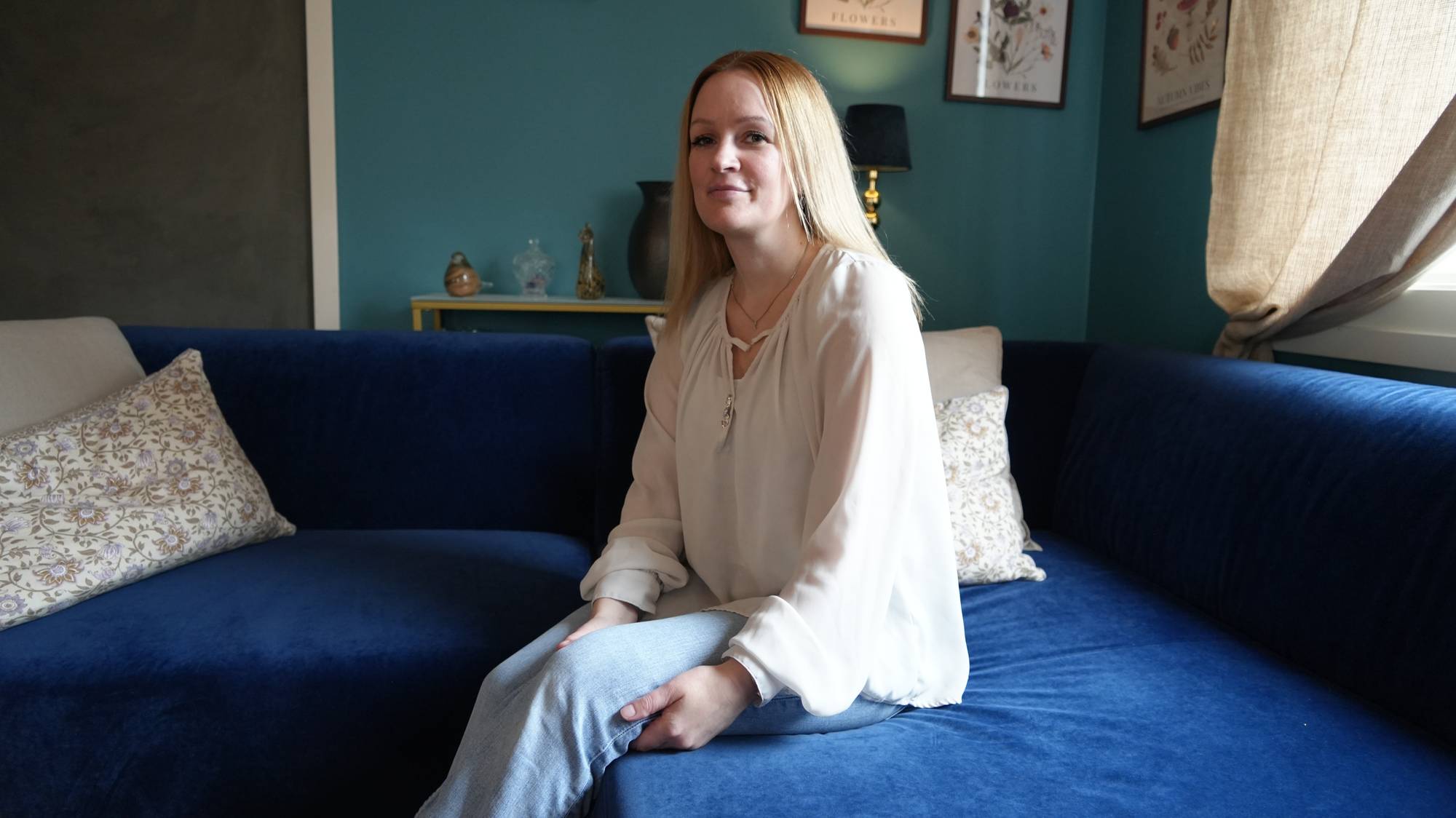 Kvinne med blondt hår sitter i en blå sofa i en stue. (Bildebeskrivelsen er laget av en KI-tjeneste)