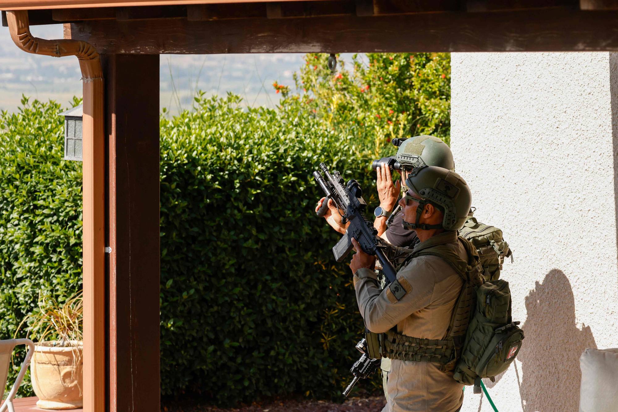 To menn i uniform med hjelm og vest er ute på et veranda. Den ene mannen holder et gevær, mens den andre ser i en kikkert. Bak dem er det grønne busker og trær. (Bildebeskrivelsen er laget av en KI-tjeneste)