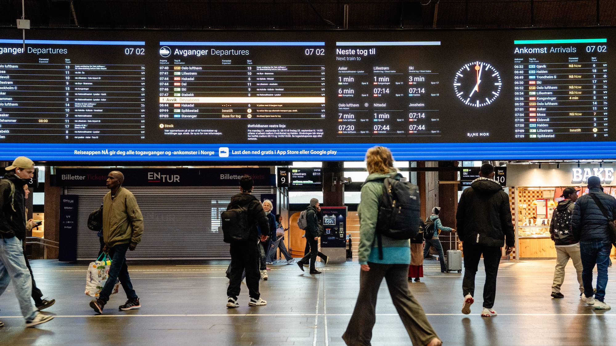 Reisende går over gulvet på en togstasjon under en stor digital informasjonstavle med togavganger. (Bildebeskrivelsen er laget av en KI-tjeneste)
