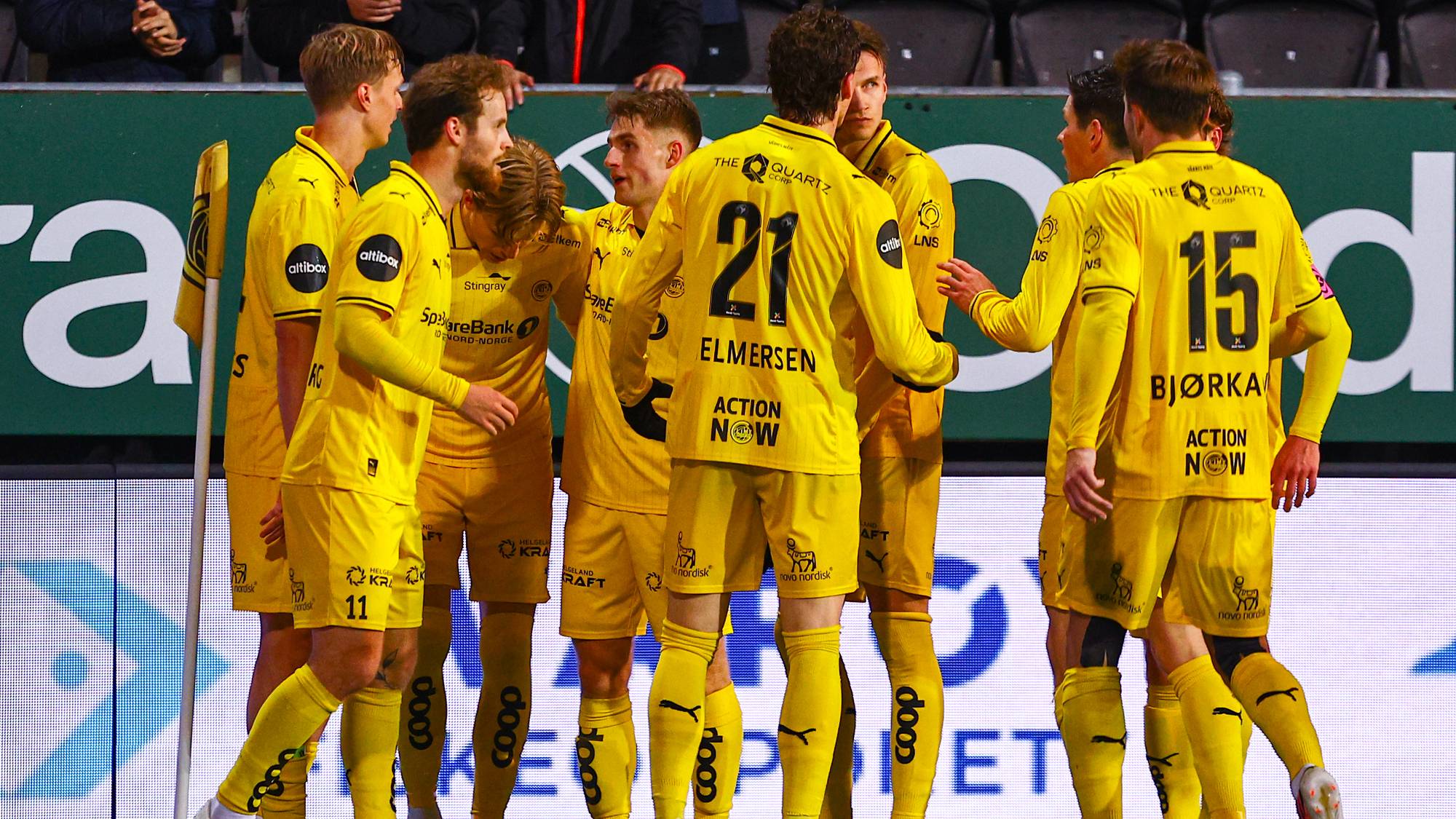 Fotballspillere i gule drakter feirer på banen foran en reklamevegg. (Bildebeskrivelsen er laget av en KI-tjeneste)