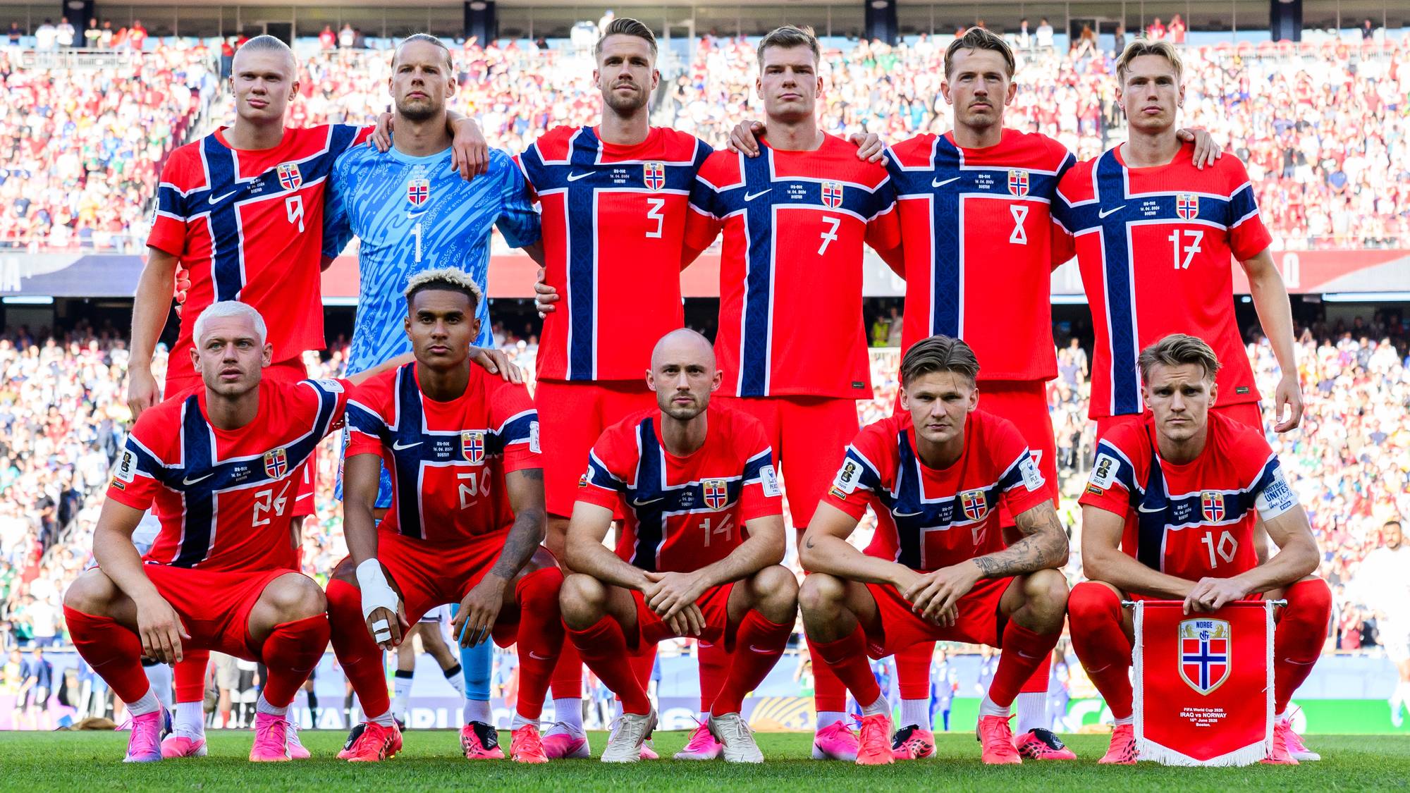 Norges herrelandslag i fotball poserer før en kamp. Spillerne er kledd i røde drakter med blått og hvitt design. Bak spillerne sitter tilskuere på tribunen. (Bildebeskrivelsen er laget av en KI-tjeneste)