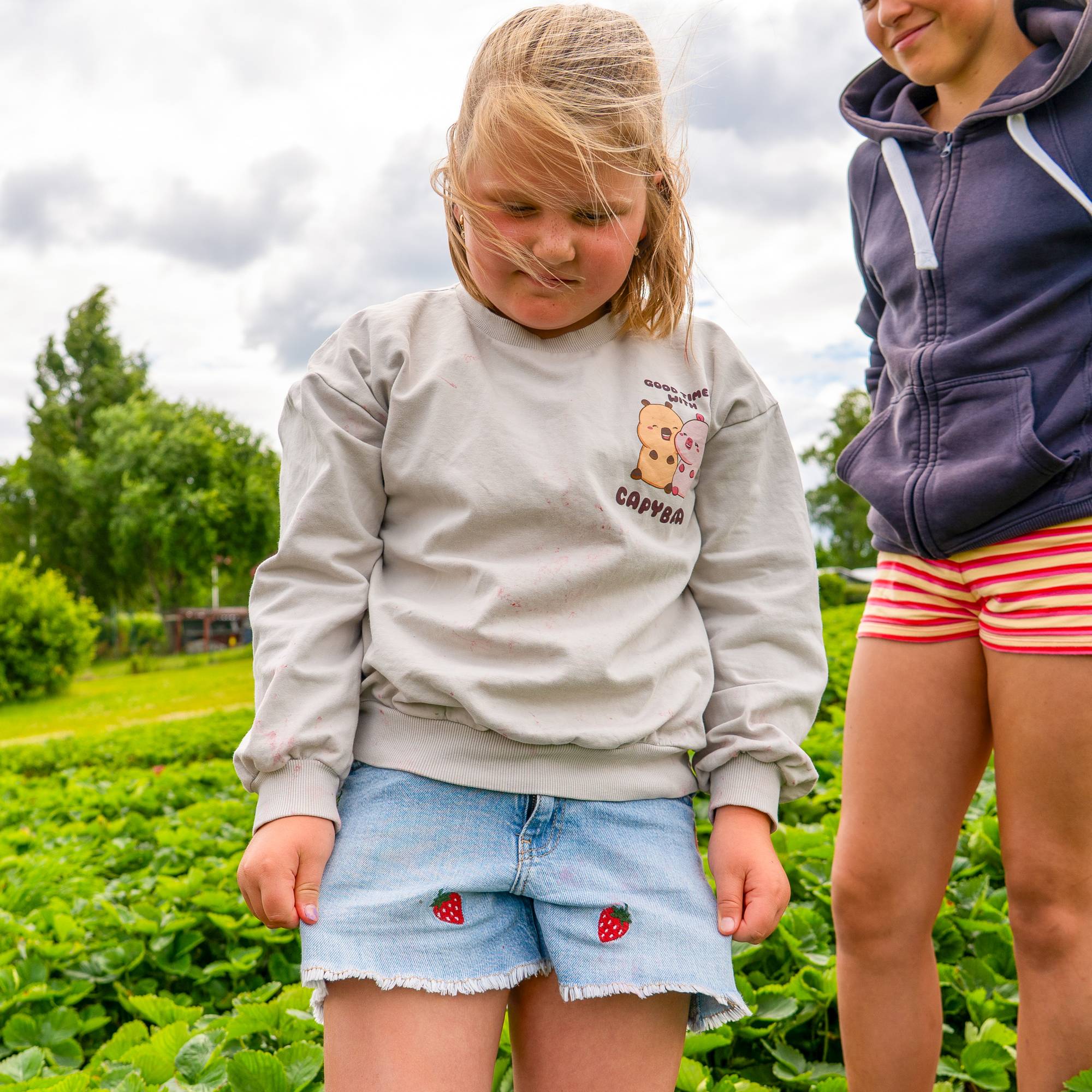 Ei jente står i jordbæråkeren og viser fram at ho har på seg ein shorts med små jordbær på. To jenter star i bakgrunnen.