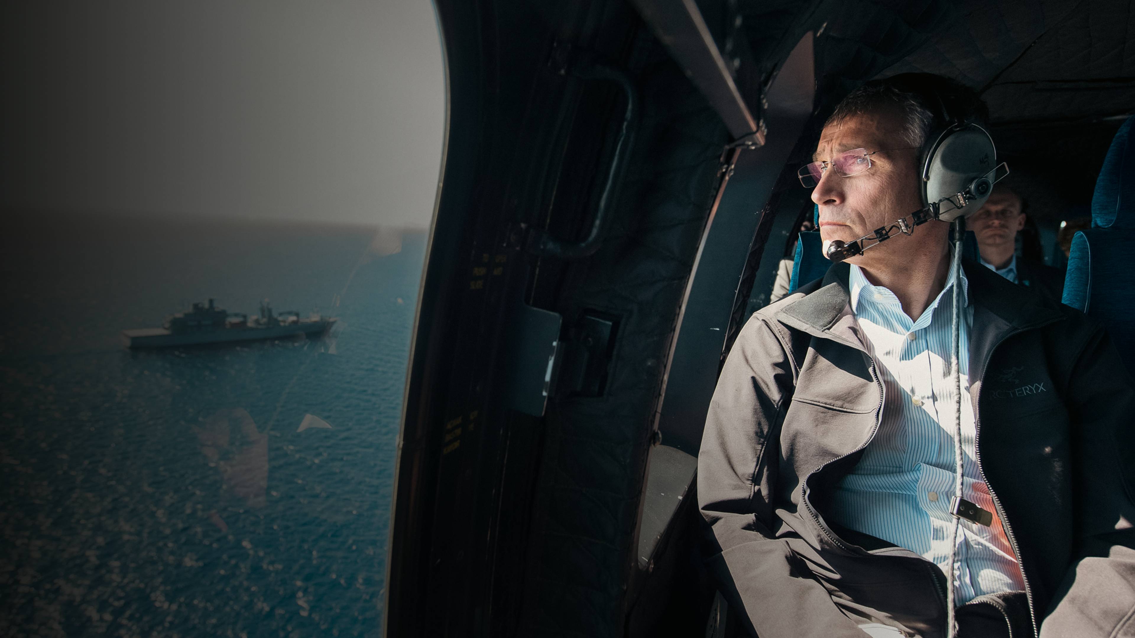 Jens Stoltenberg sitter i et helikopter og ser ned på havet. Under kan man se et skip.