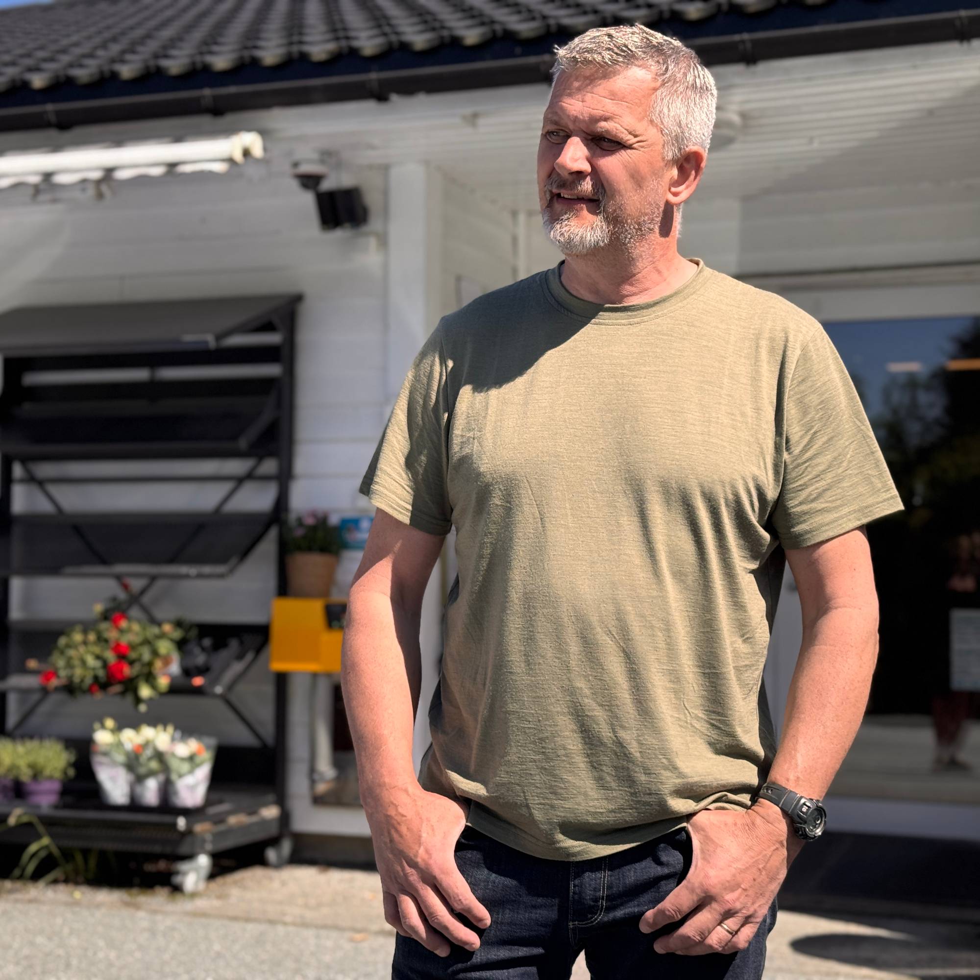 En mann står foran en hvit bygning med en takhøyde. Mannen har grått hår og skjegg og er kledd i en olivenfarget t-skjorte og mørke bukser. Bak ham er det en blomsterhylle med planter og en blå bil. (Bildebeskrivelsen er laget av en KI-tjeneste)