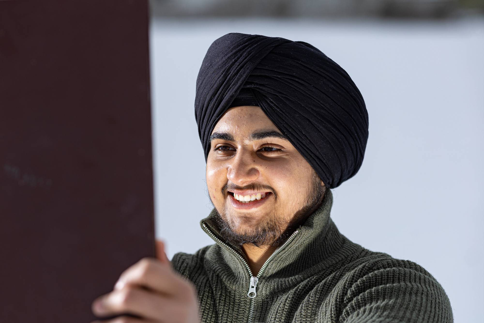 Et nærbilde av Gurveer med sin sort turban og grønn genser. Det er snø i bakgrunnen. Han smiler stort. Han holder noe i hendene som han ser på, men vi ser ikke hva han holder. Er det et bilde? Et speil?