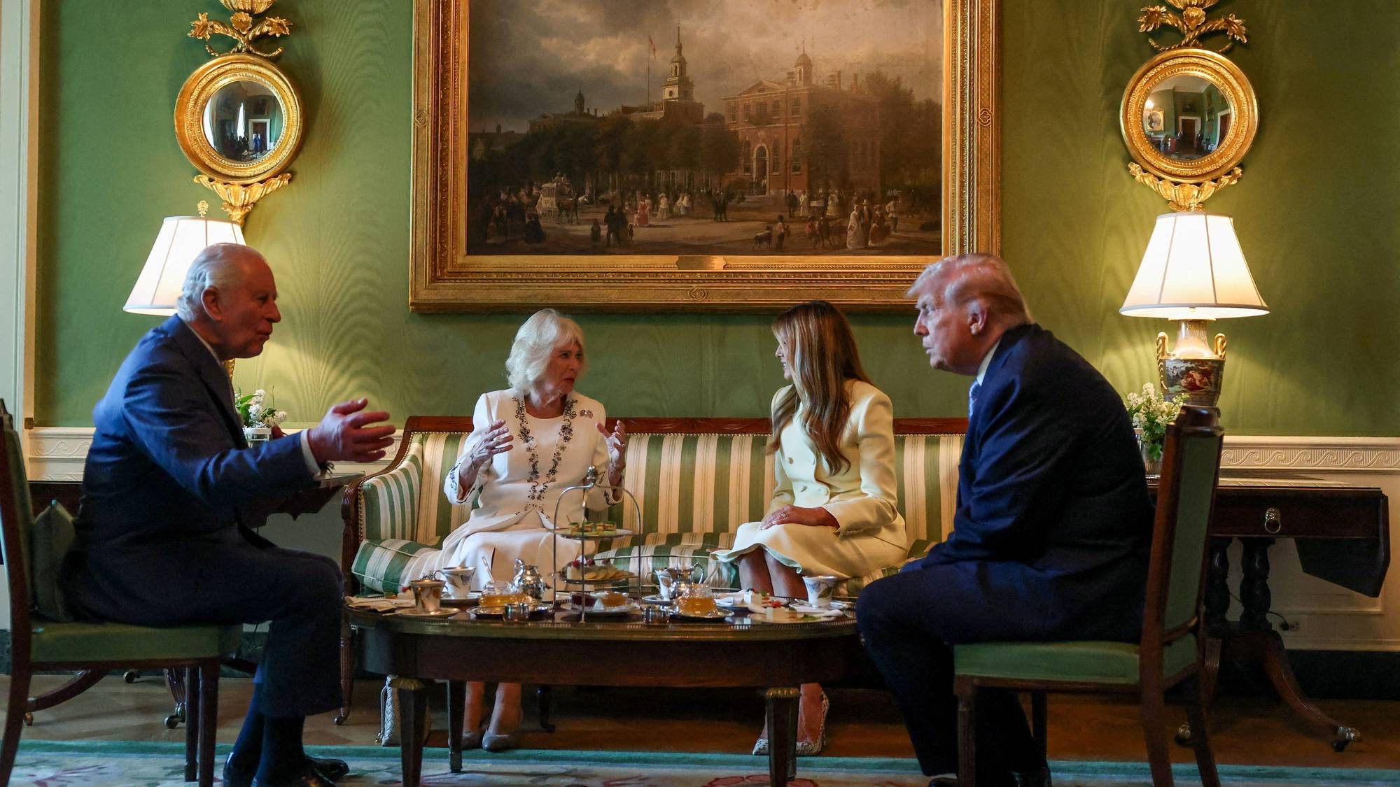 King Charles III, Queen Camilla and Donald Trump meet for tea. They are seated in a room with green walls and a large painting. Tea is served on a table between them. (Bildebeskrivelsen er laget av en KI-tjeneste)
