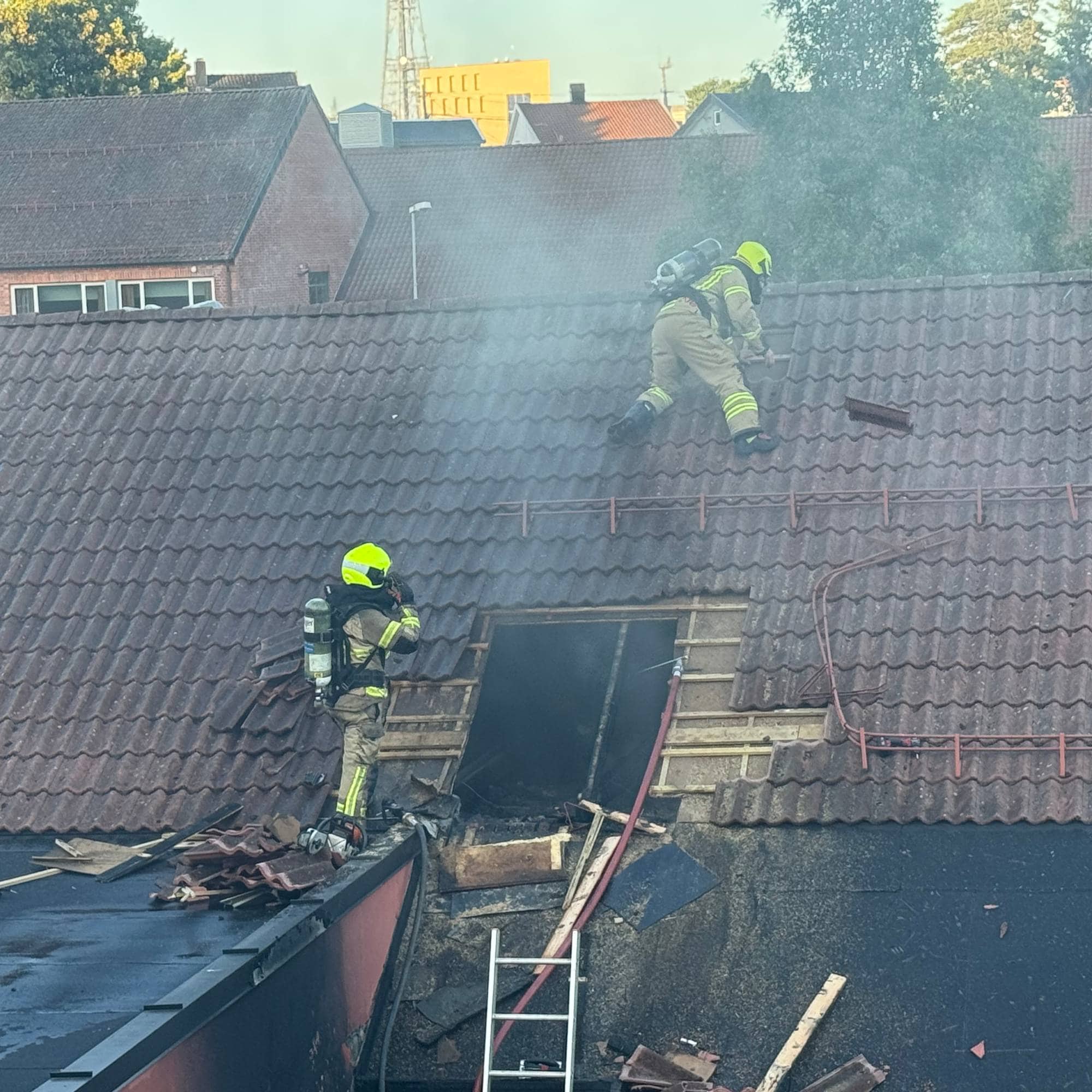 Brannmannskaper i fullt utstyr arbeider på et hustak med et åpent hull. Røyk stiger opp fra taket. (Bildebeskrivelsen er laget av en KI-tjeneste)