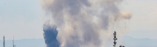 En stor røyksøyle stiger opp over et bybilde. Det er en skyggefull kjerne i røyken. Antenner og bygninger er synlige i forgrunnen. (Bildebeskrivelsen er laget av en KI-tjeneste)