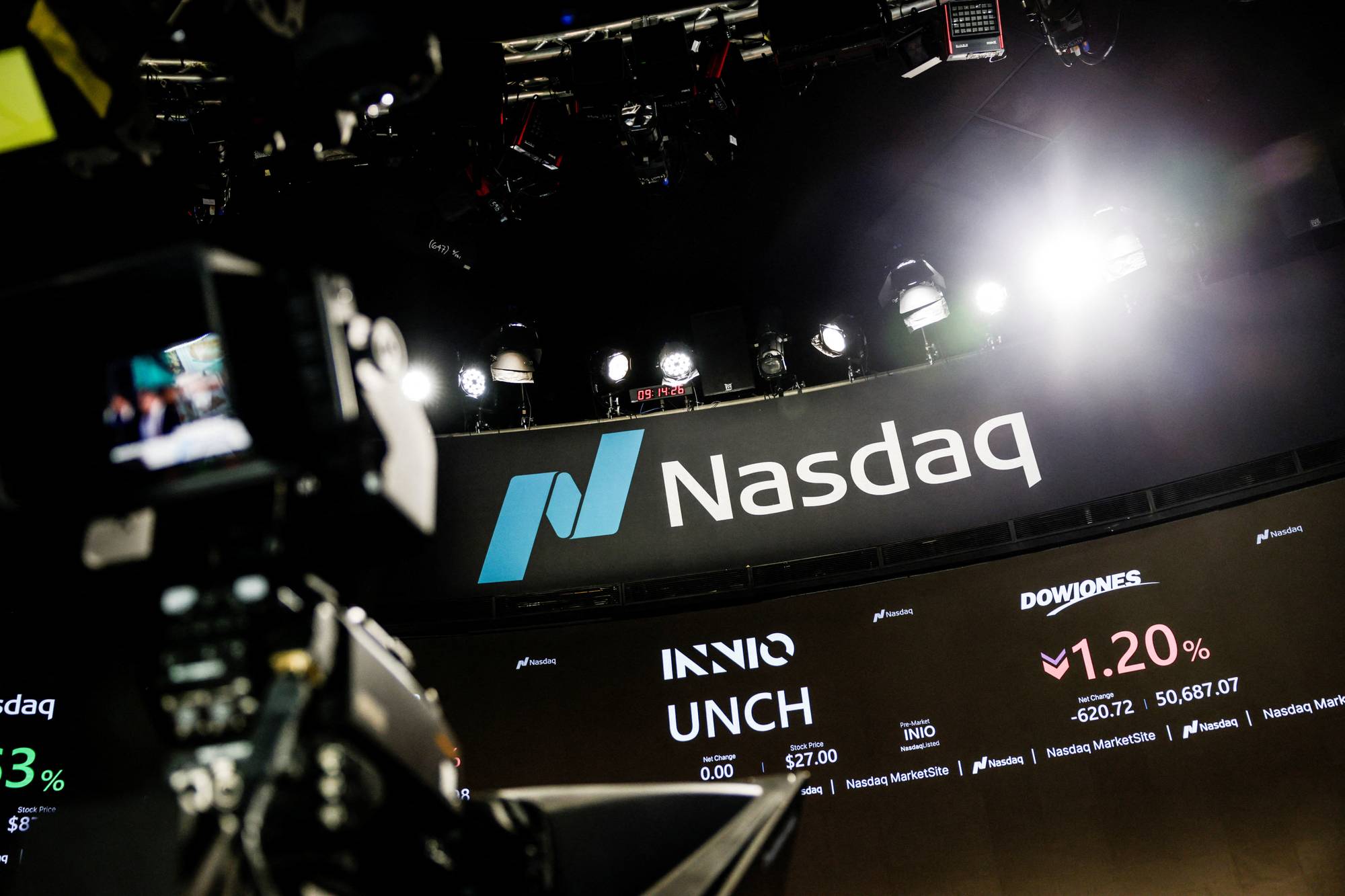 Stor skjerm viser Nasdaq-logo og aksjekurser. En TV-skjerm viser et bilde av personer, og en TV-monitor er synlig i forgrunnen. Flere spotlykter er rettet mot scenen. (Bildebeskrivelsen er laget av en KI-tjeneste)