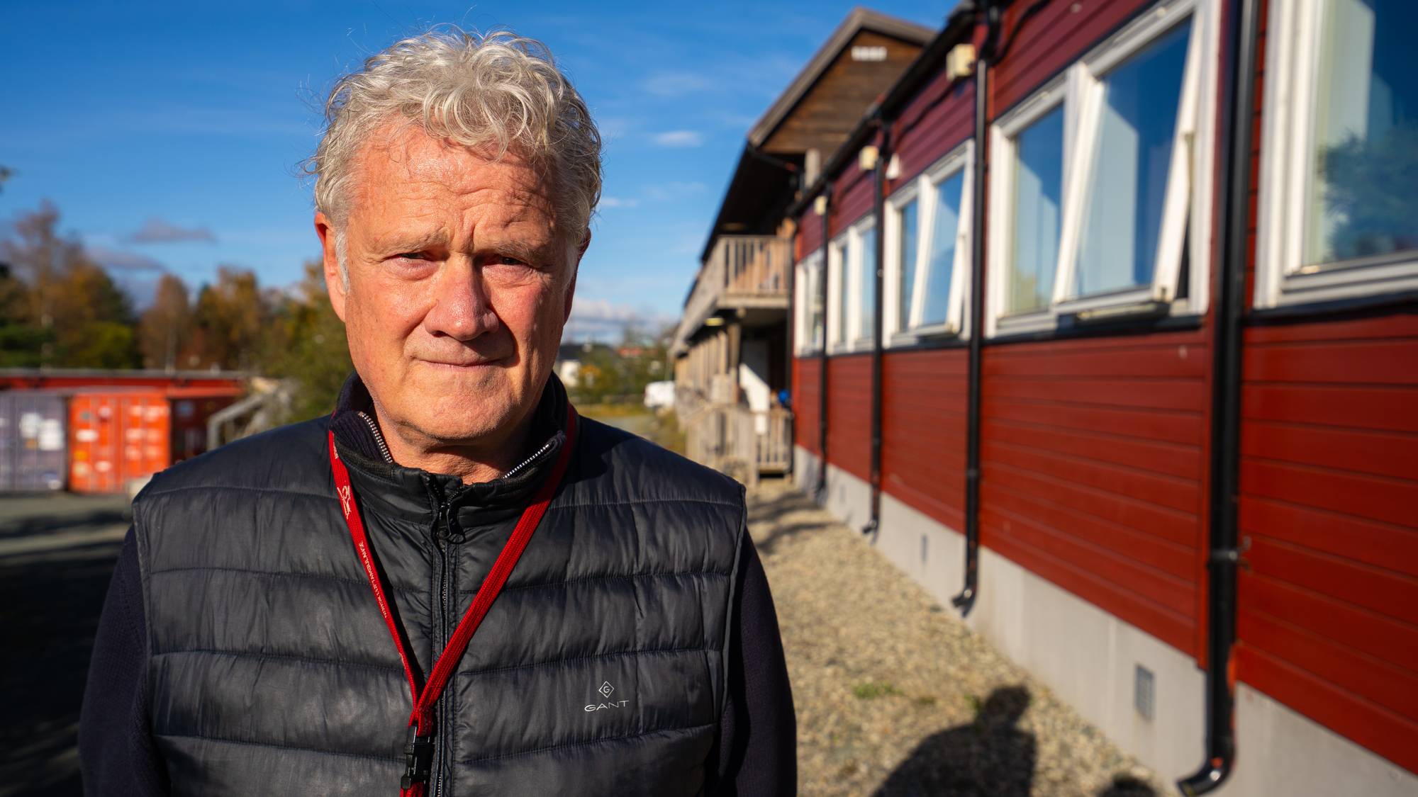 En eldre mann med grått hår står utendørs foran en rød trebygning. Han har på seg en svart vest. (Bildebeskrivelsen er laget av en KI-tjeneste)