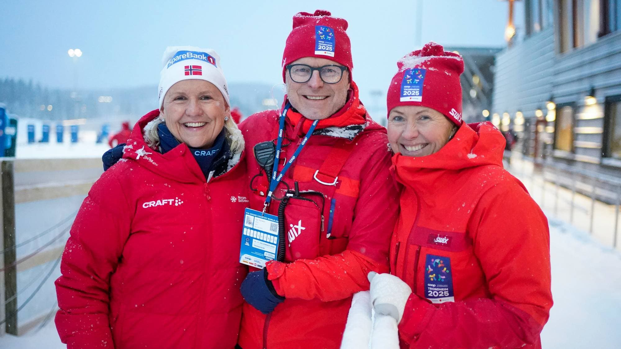 Tre personer i røde jakker og luer smiler. Den midterste personen har briller og ID-kort. Bak dem er snødekte omgivelser med bygninger og lys. (Bildebeskrivelsen er laget av en KI-tjeneste)