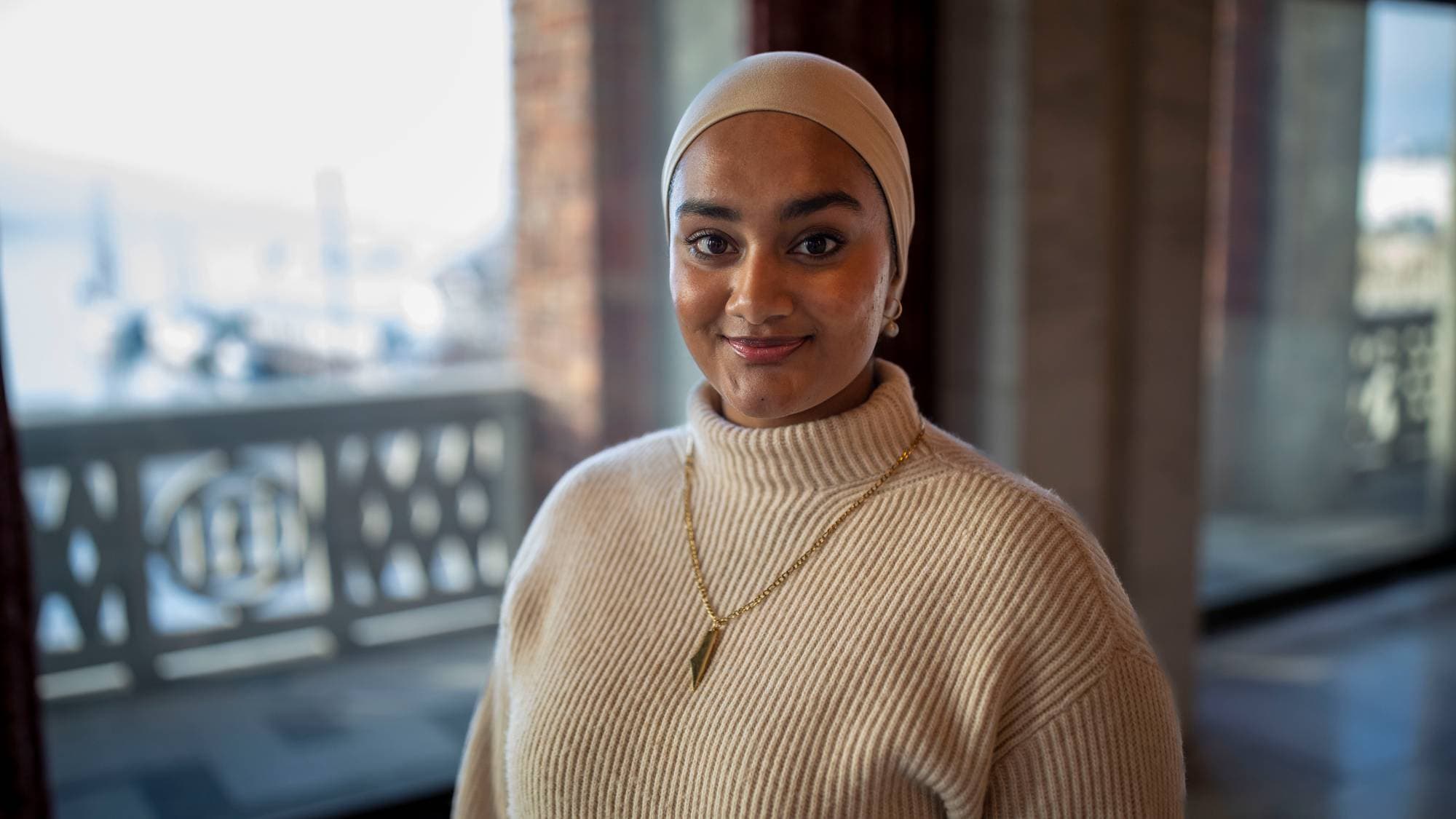 Portrett av en smilende kvinne i lys hijab og strikkegenser med smykke innendørs. (Bildebeskrivelsen er laget av en KI-tjeneste)