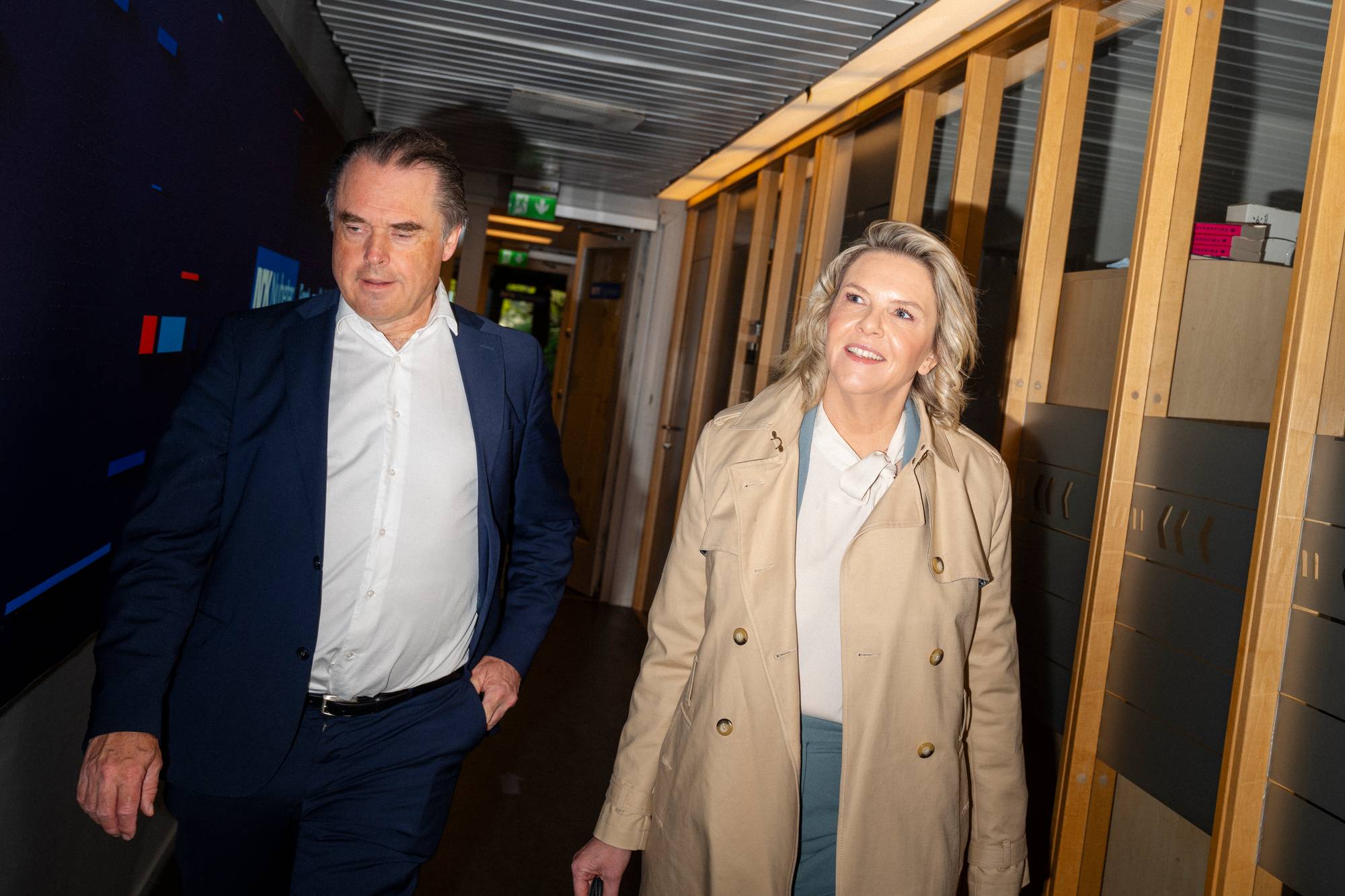En mann i mørk dress og en kvinne i beige frakk går sammen i en korridor. Kvinnen smiler mens mannen ser ned. I bakgrunnen sees en mørk vegg med blå og røde former, samt en vegg med vertikale trepaneler. (Bildebeskrivelsen er laget av en KI-tjeneste)