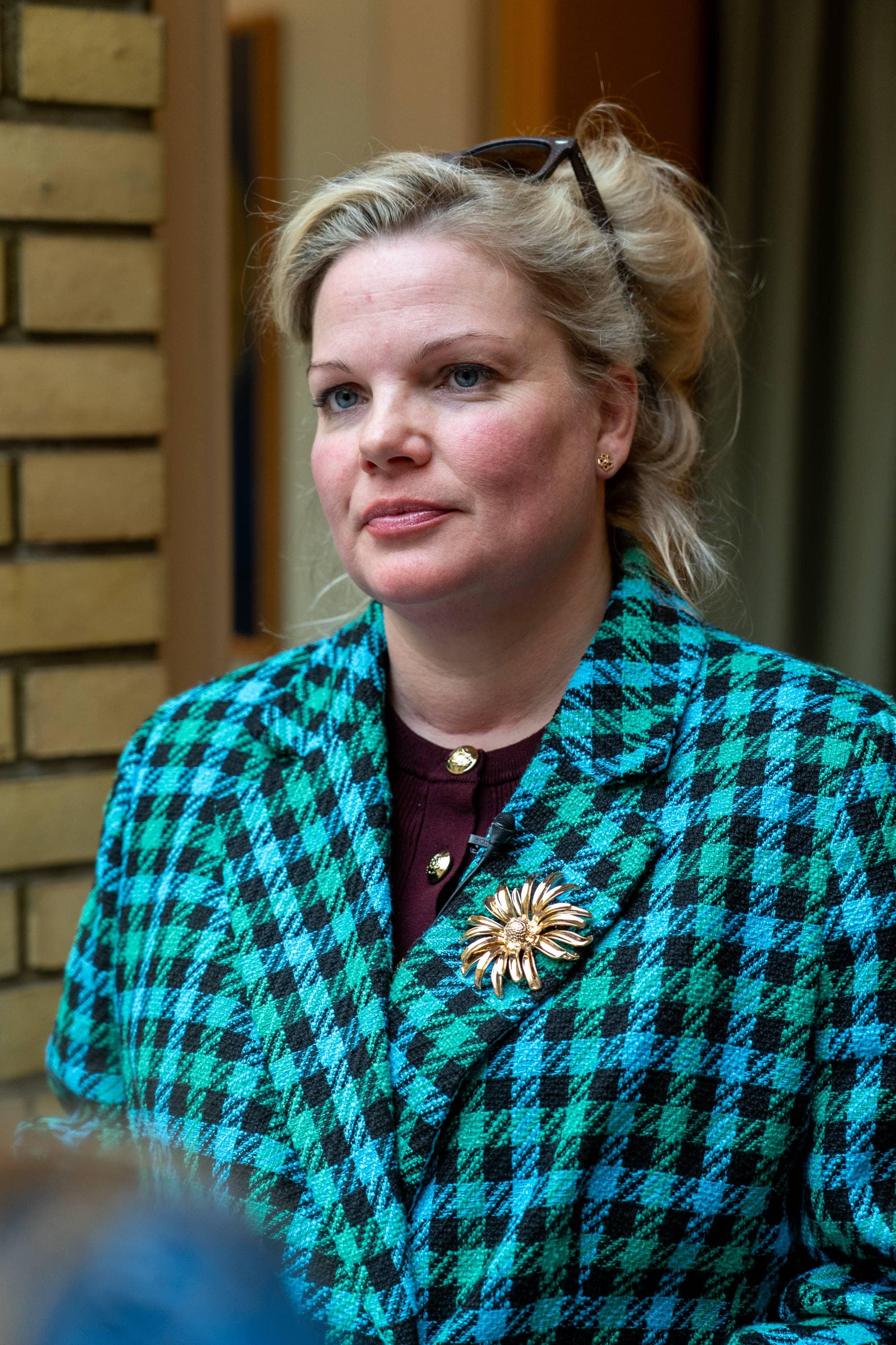 En kvinne med blondt hår og solbriller på hodet. Hun har på seg en rutete jakke i blått og grønt med en brosje. Kvinnen står foran en mursteinsvegg og en gardin. (Bildebeskrivelsen er laget av en KI-tjeneste)