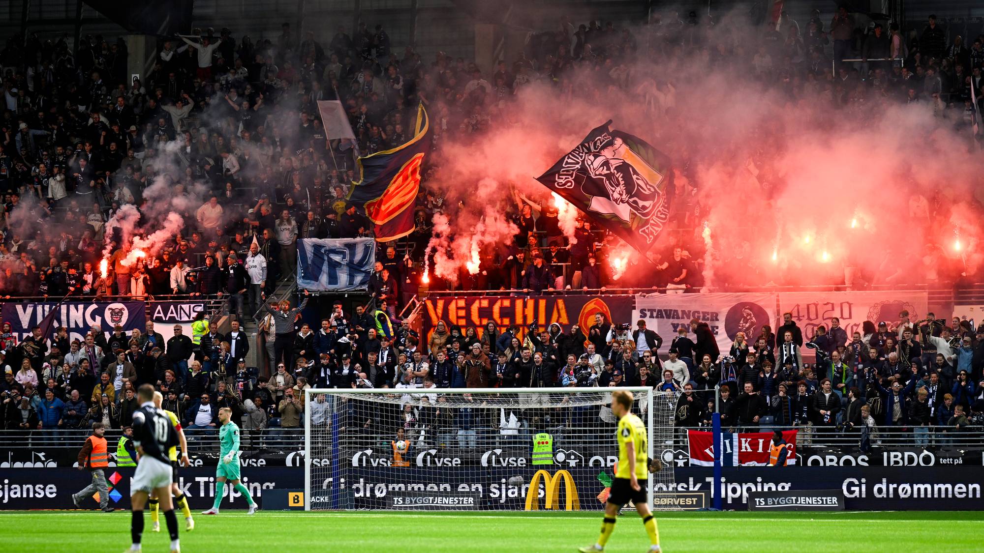 Fansen tenner pyroteknikk på en fotballkamp. Tilskuere sitter på tribunen og ser på. En stor mengde røyk fyller luften over tribunene. Spillerne er på gressmatten. (Bildebeskrivelsen er laget av en KI-tjeneste)