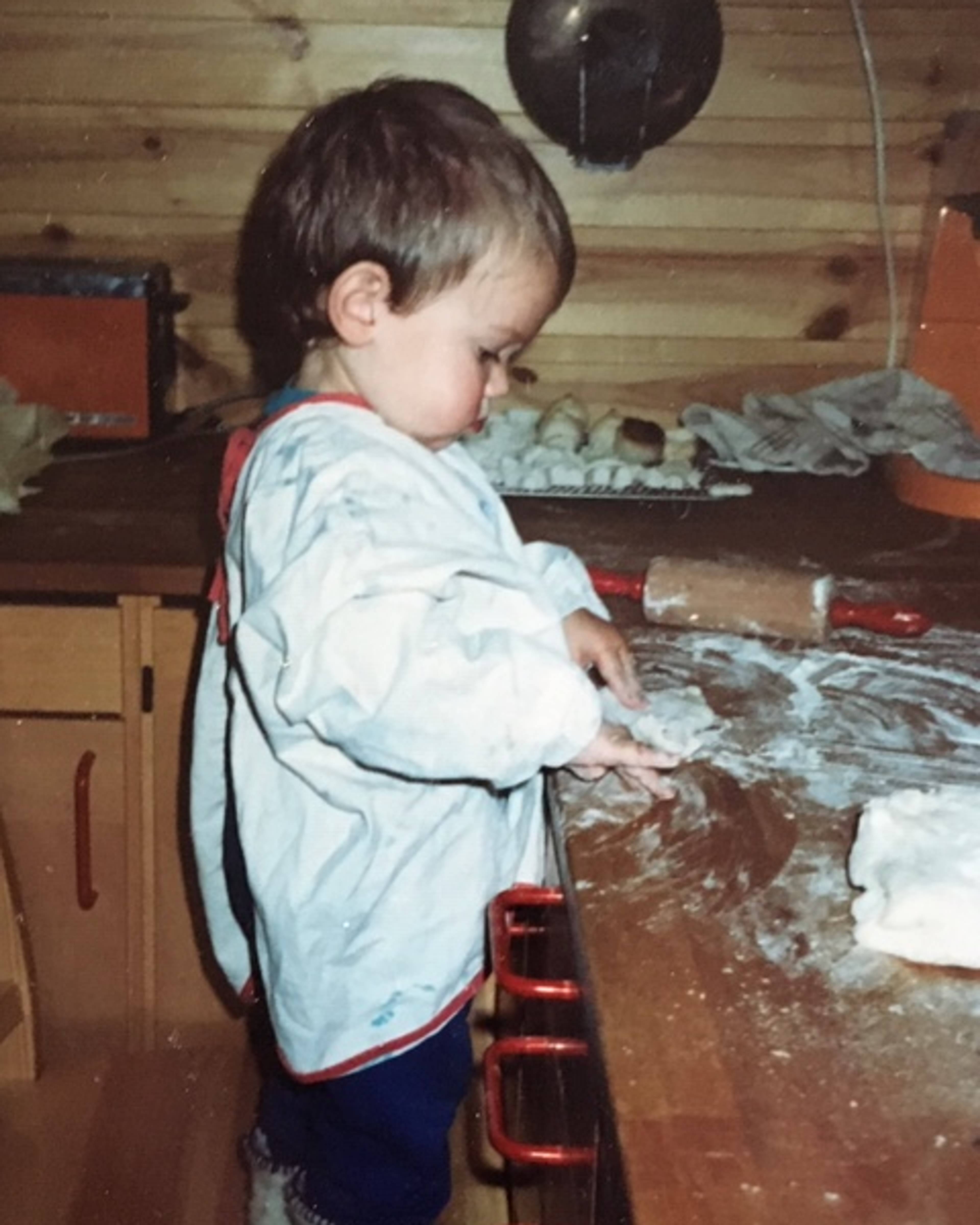 Et lite barn med smekke leker med mel på en kjøkkenbenk. Barnet står ved siden av en kjevle. Kjøkkenet har tømmervegger. (Bildebeskrivelsen er laget av en KI-tjeneste)