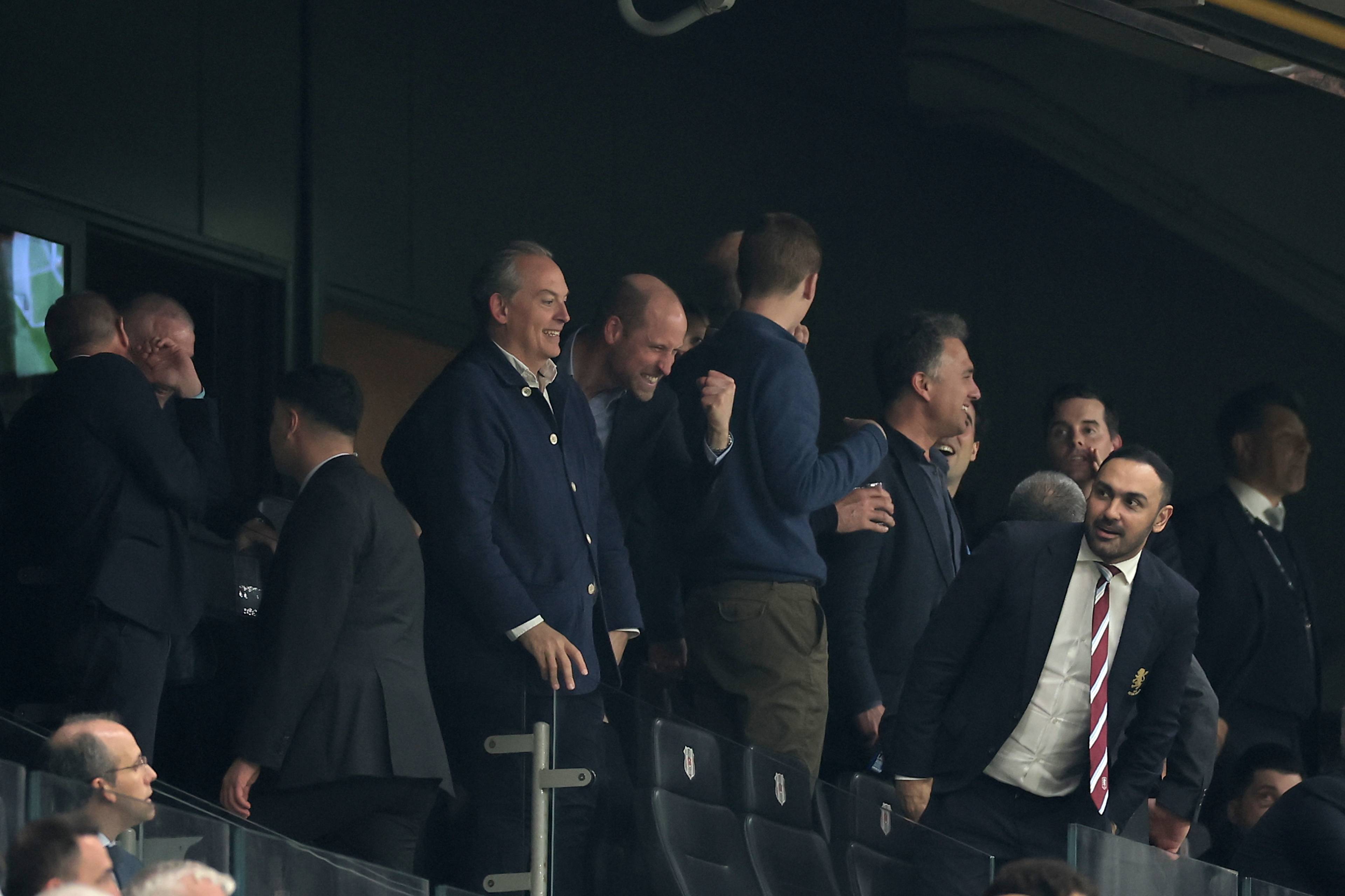 En gruppe menn i mørke dresser og jakker står og ser utover. Noen snakker sammen, mens andre ser mot høyre. De befinner seg på tribunen på en idrettsarena. Bildet har et vannmerke fra NTB. (Bildebeskrivelsen er laget av en KI-tjeneste)