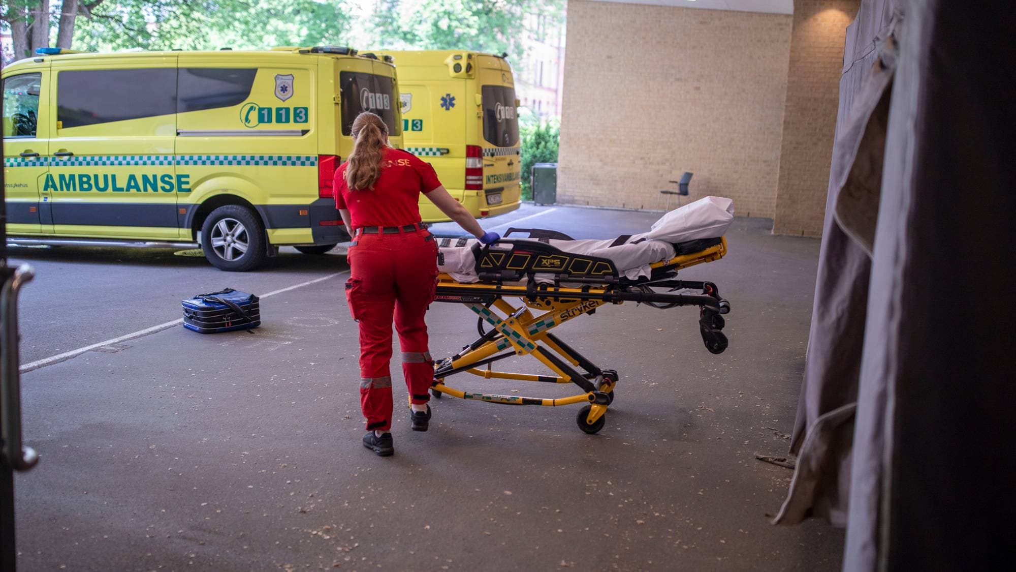 En person i røde arbeidsklær håndterer en ambulanse-tralle. En gul ambulanse står parkert bak. Omgivelsene er en asfaltert uteplass med en mursteinsbygning i bakgrunnen. (Bildebeskrivelsen er laget av en KI-tjeneste)