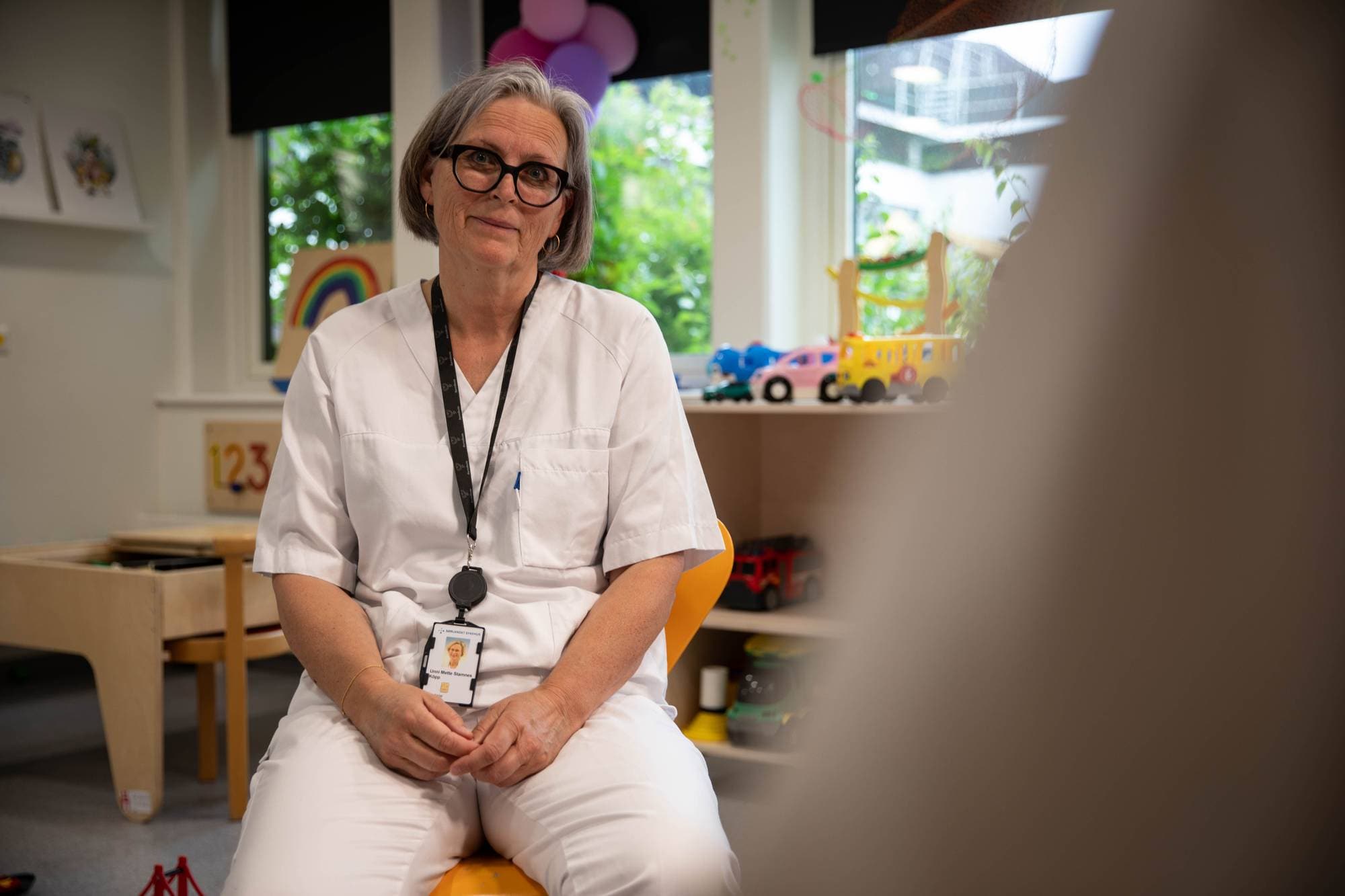 En kvinne i hvite arbeidsklær sitter og ser mot kamera. Hun har briller og et ID-kort hengende rundt halsen. Bak henne er det leker og en regnbue-illustrasjon på veggen. Rommet ser ut som et barnehagerom eller et lignende sted for barn. (Bildebeskrivelsen er laget av en KI-tjeneste)