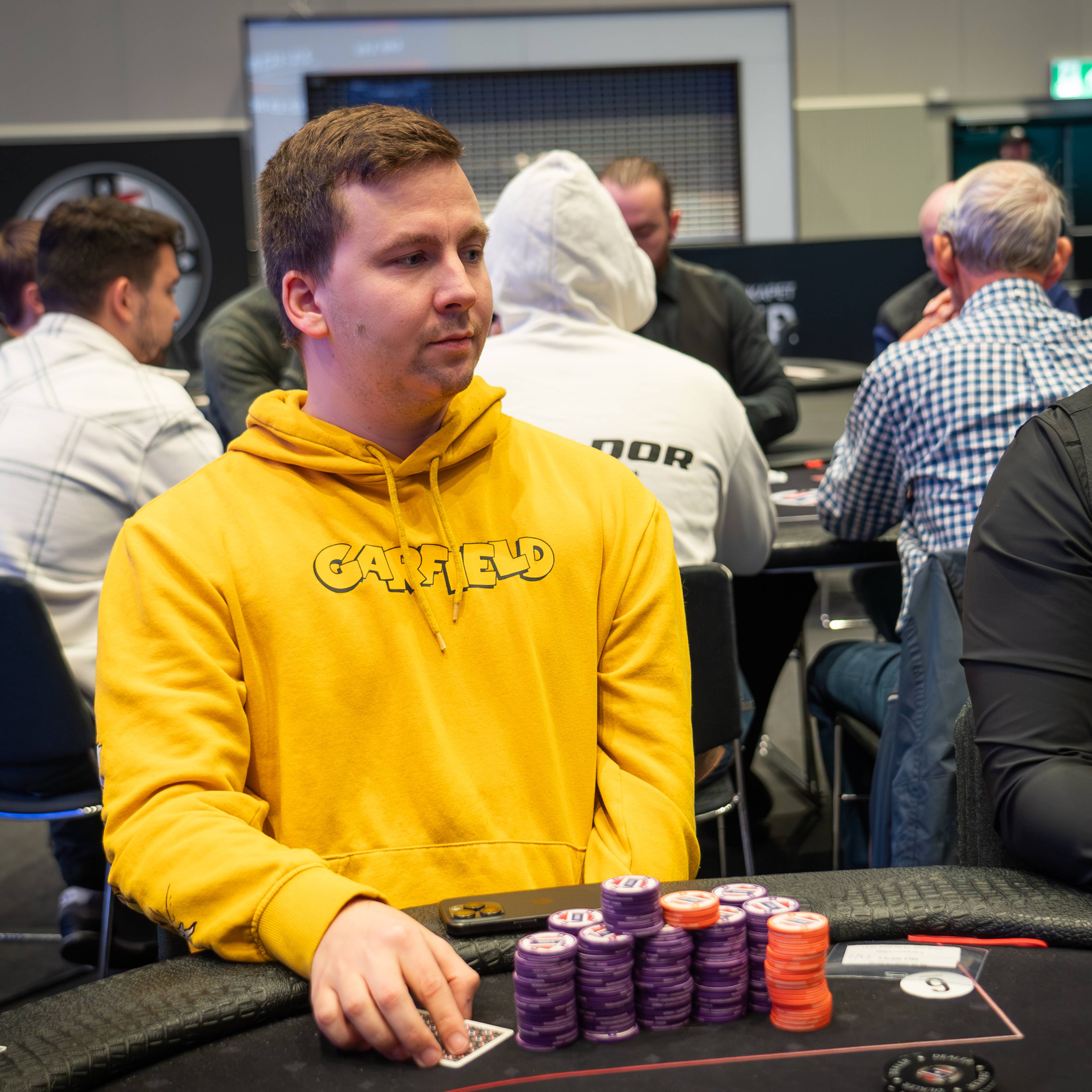Spiller i gul hettegenser ved et pokerbord med sjetonger foran seg i en turnering.