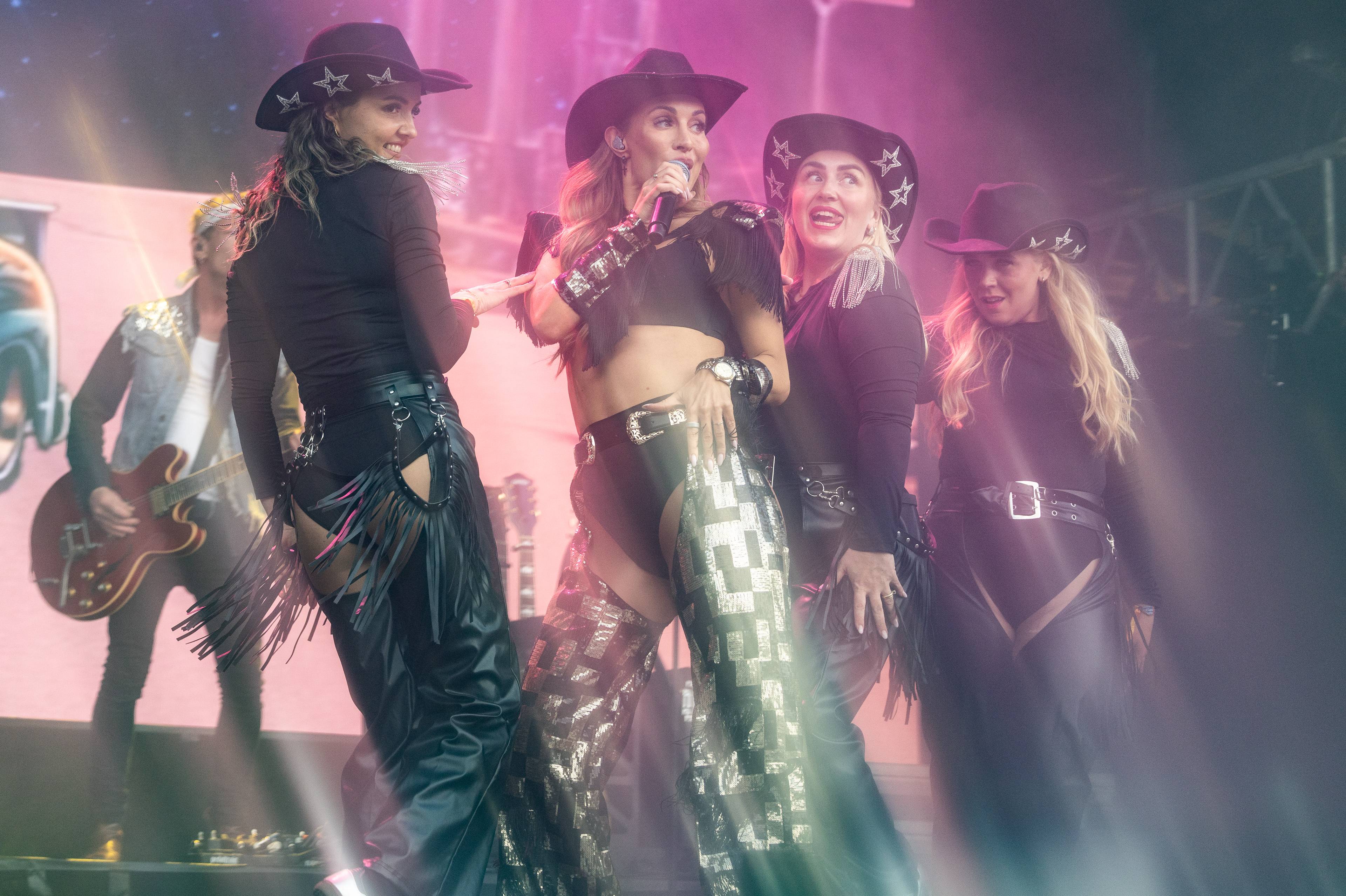 En gruppe kvinnelige artister opptrer på en scene iført cowboyhatter og scenekostymer. En av artistene holder en mikrofon og synger. En mann spiller gitar i bakgrunnen. Lysstråler og tåke skaper en atmosfære på scenen. (Bildebeskrivelsen er laget av en KI-tjeneste)