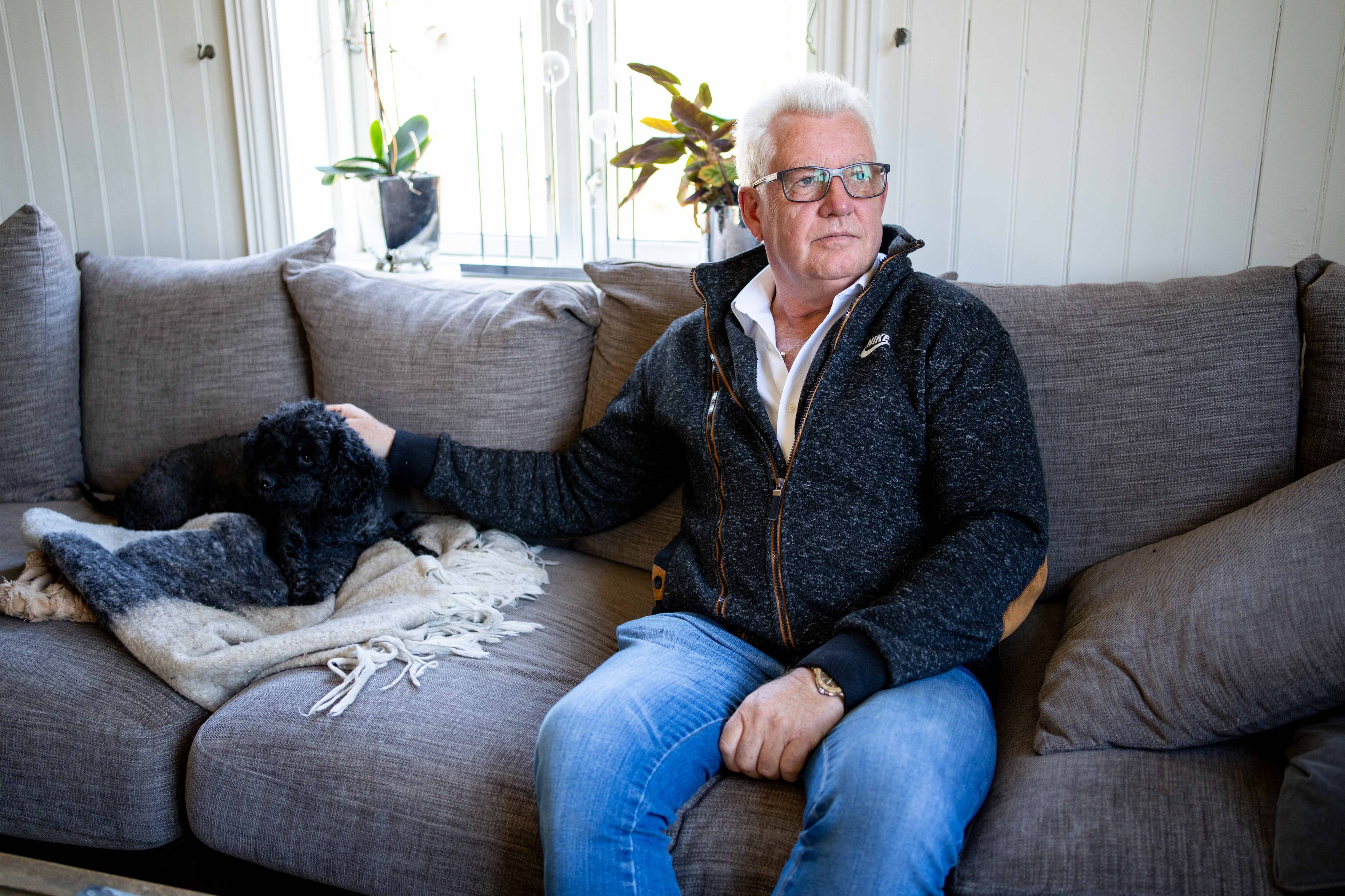 Erik Arntzen sitter i sofaen med hunden Lillevenn. Han ser ut til siden. Han har hvitt hår og briller.