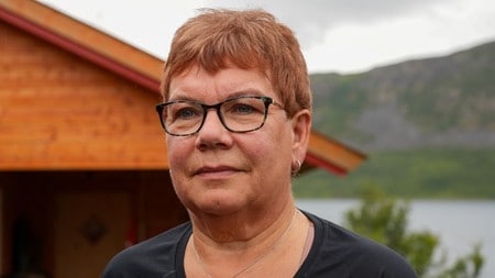 Kvinne med briller og kort rødt hår står foran en hytte ved et vann og fjell. (Bildebeskrivelsen er laget av en KI-tjeneste)