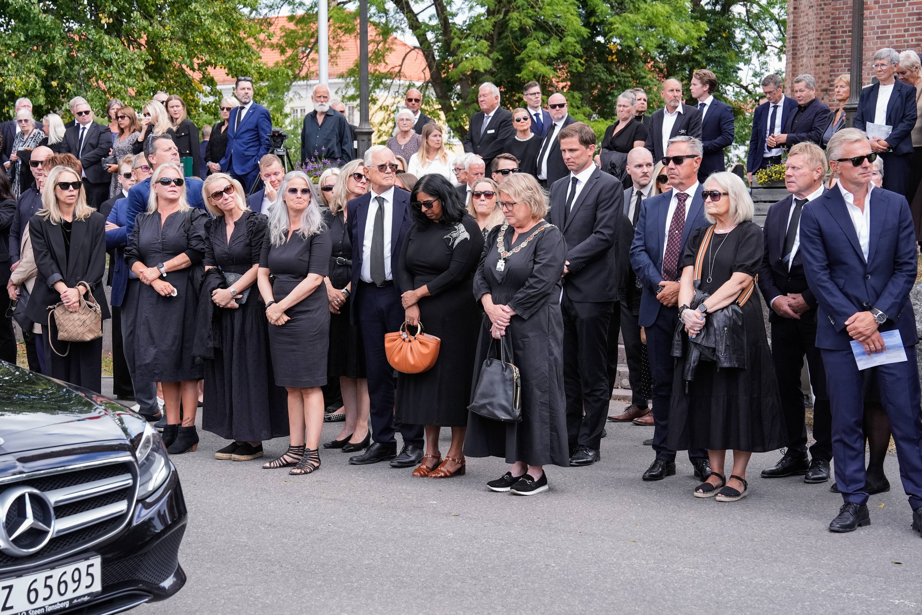 En folkemengde i mørke klær står samlet utendørs, delvis foran en mørk bil.