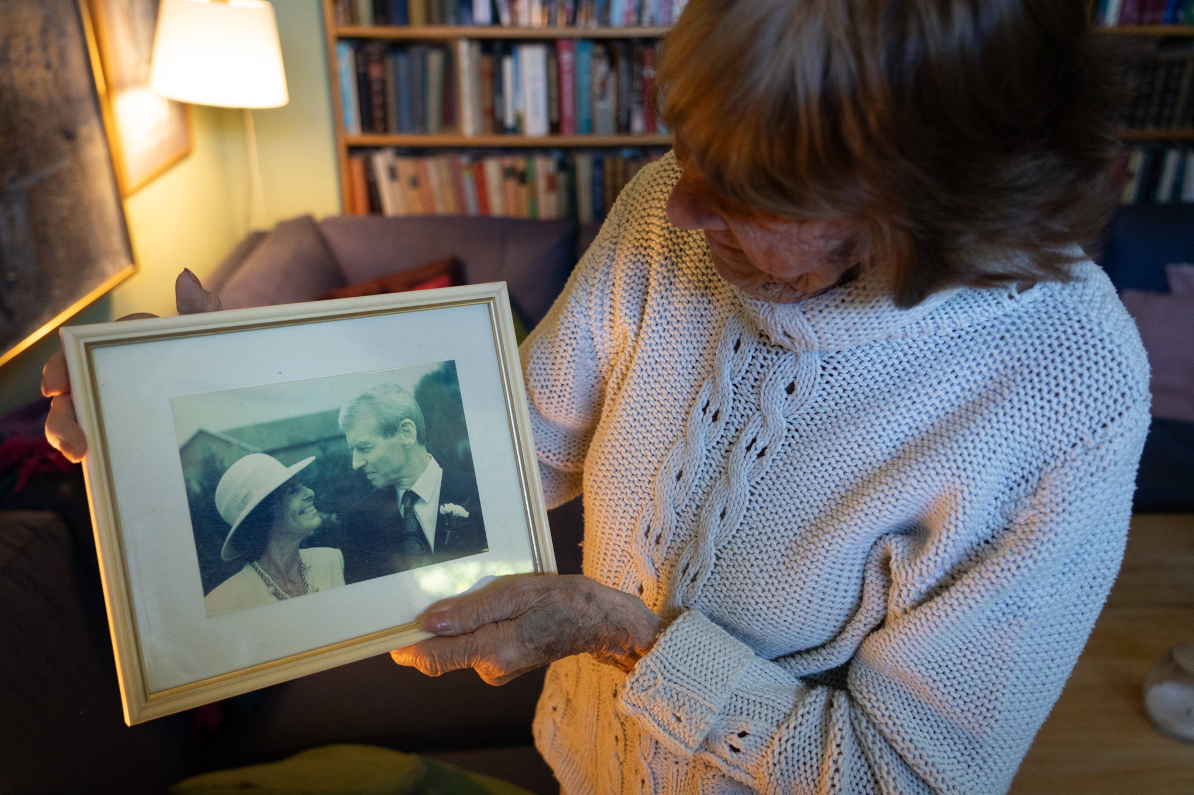 En eldre kvinne holder et innrammet bilde av et brudepar. Bildet viser en kvinne med hatt og en mann i dress som ser på hverandre. De er innenfor en bokhylle. (Bildebeskrivelsen er laget av en KI-tjeneste)