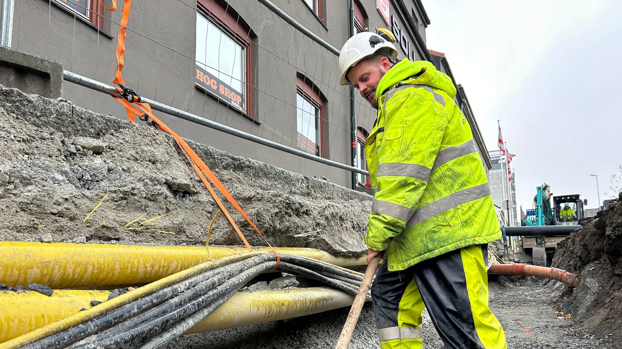 En arbeider i gul refleksdress og hjelm graver med en spade i en grøft. Gul isolasjon og kabler ligger i grøften. I bakgrunnen sees en gravemaskin og en bygning. Det er et utendørs bilde. (Bildebeskrivelsen er laget av en KI-tjeneste)