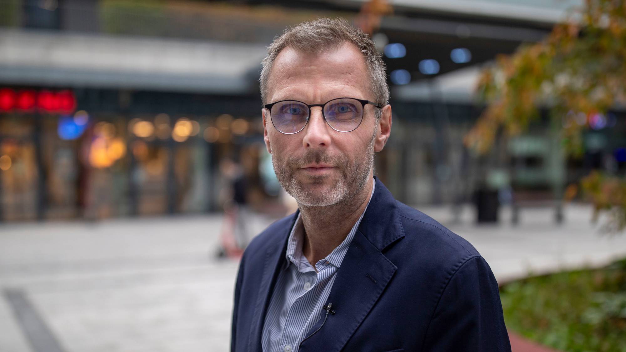 Portrett av mann i 40-årene med briller og mørk dressjakke, stående utendørs foran en bygning. (Bildebeskrivelsen er laget av en KI-tjeneste)