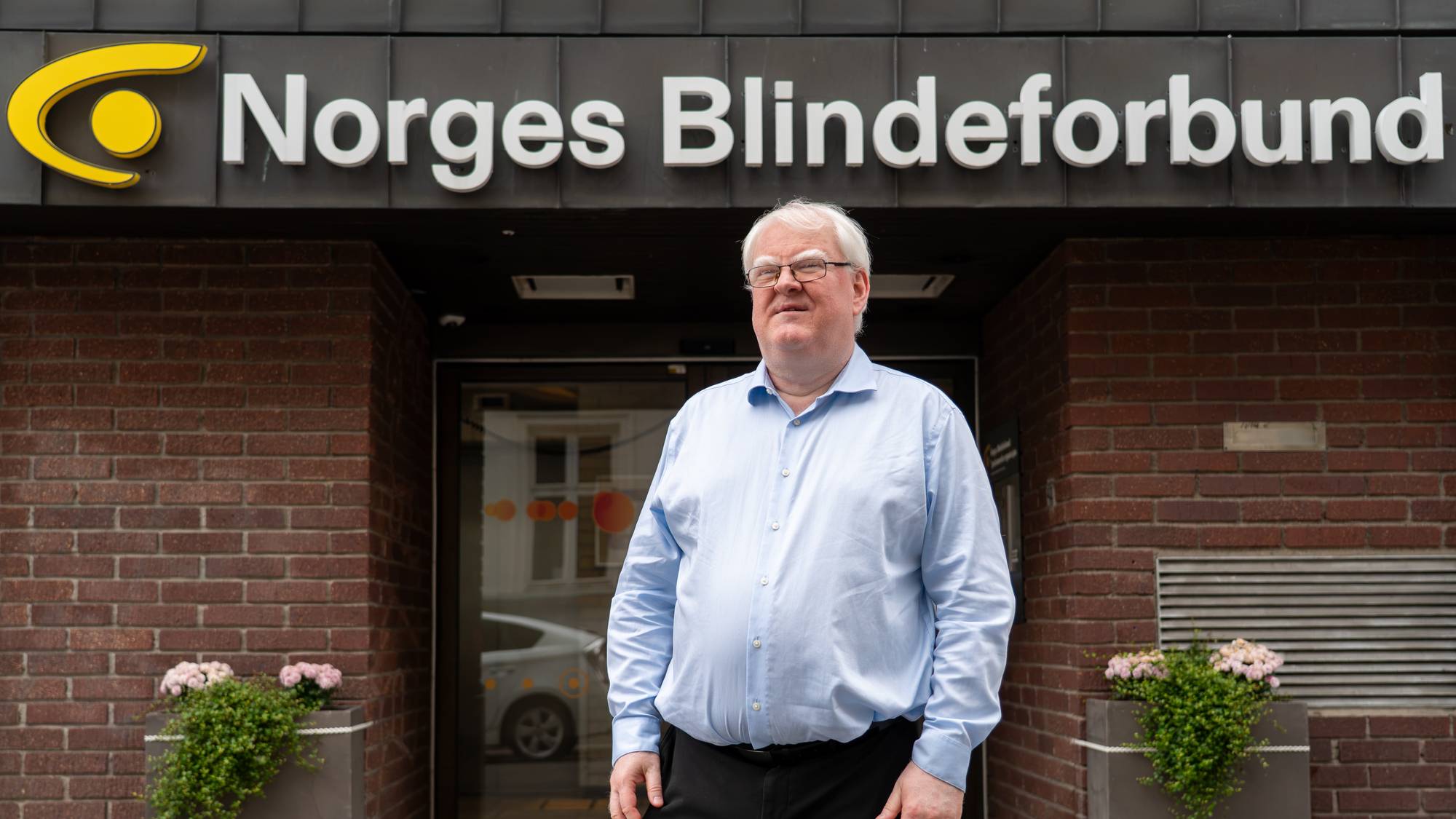 Mann med briller står foran inngangen til Norges Blindeforbunds lokaler. (Bildebeskrivelsen er laget av en KI-tjeneste)