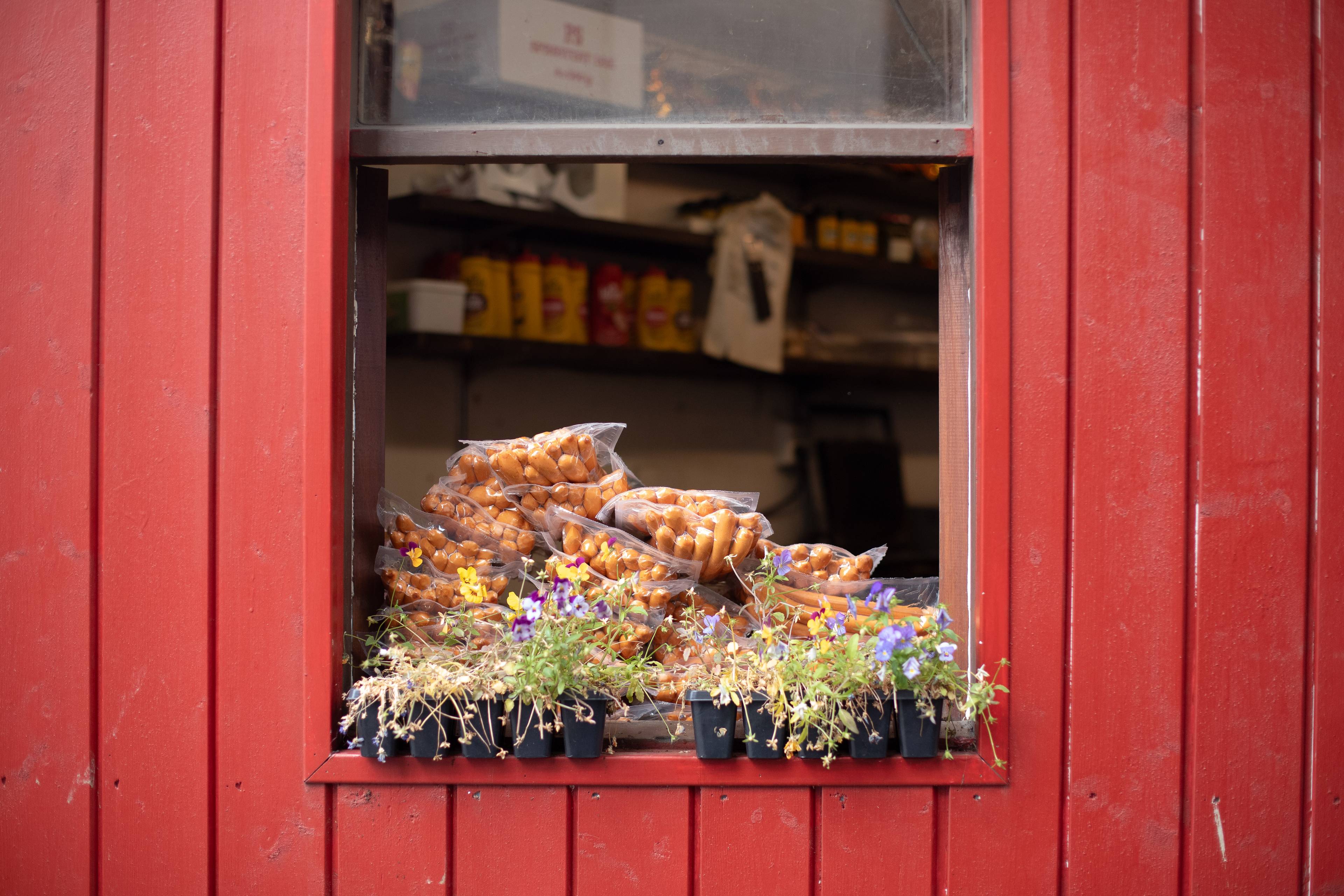 en haug med pølser ligger i en vinduskarm, bygget er rødt i tre og foran pølsene ligger det blomster