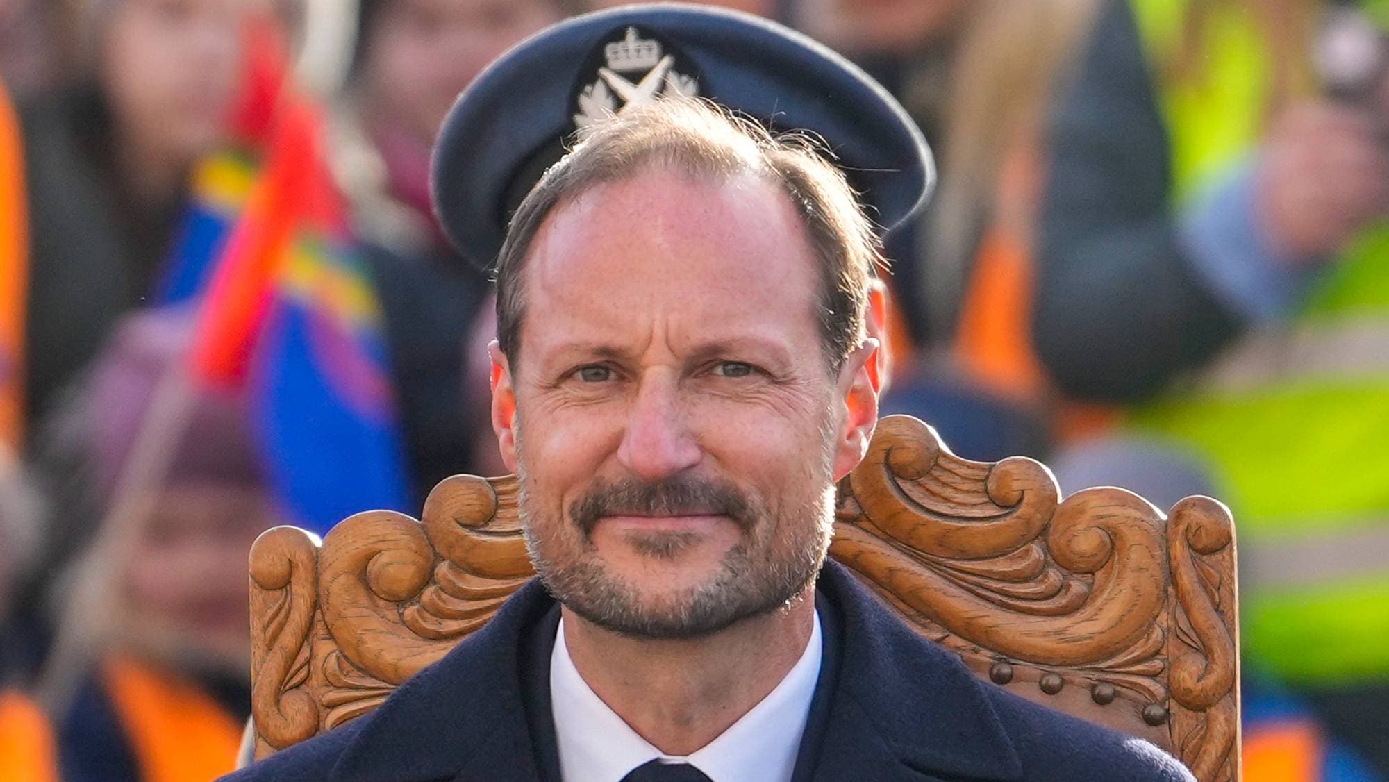 Kronprins Haakon sitter i en utskåret trestol iført uniformslue og mørk dress. Bakgrunnen er uklar. (Bildebeskrivelsen er laget av en KI-tjeneste)
