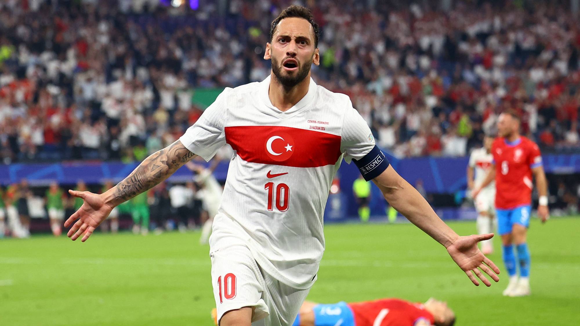 En tyrkisk fotballspiller med nummer 10 jubler med armene utstrakt. En annen spiller ligger på bakken. En stor folkemengde sitter på tribunen i bakgrunnen. (Bildebeskrivelsen er laget av en KI-tjeneste)