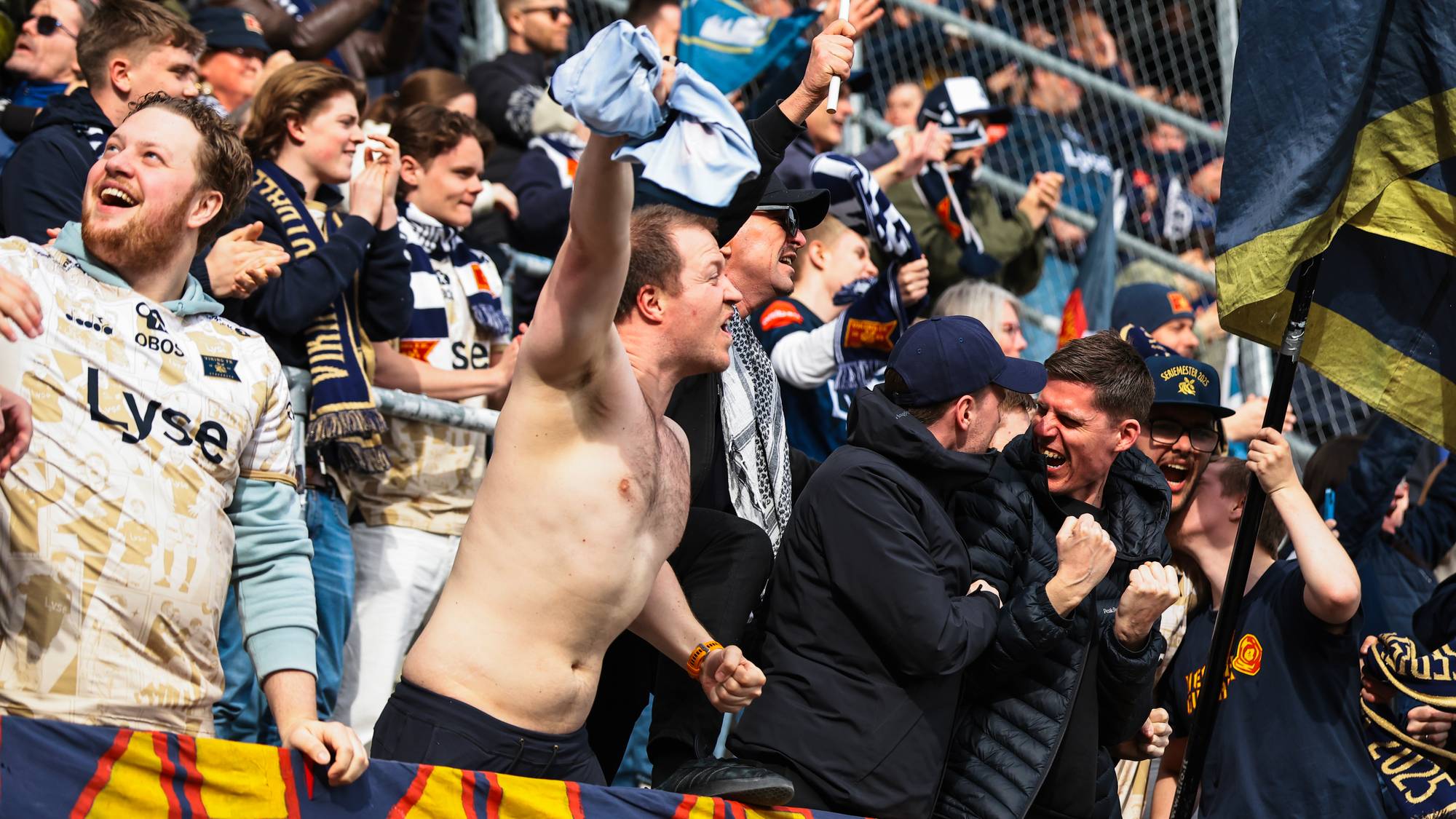 En bar overkropp mann jubler og holder opp et lyst tøy. Tilskuere med supporterutstyr i bakgrunnen. En stor fane med gult og blått mønster er synlig. (Bildebeskrivelsen er laget av en KI-tjeneste)