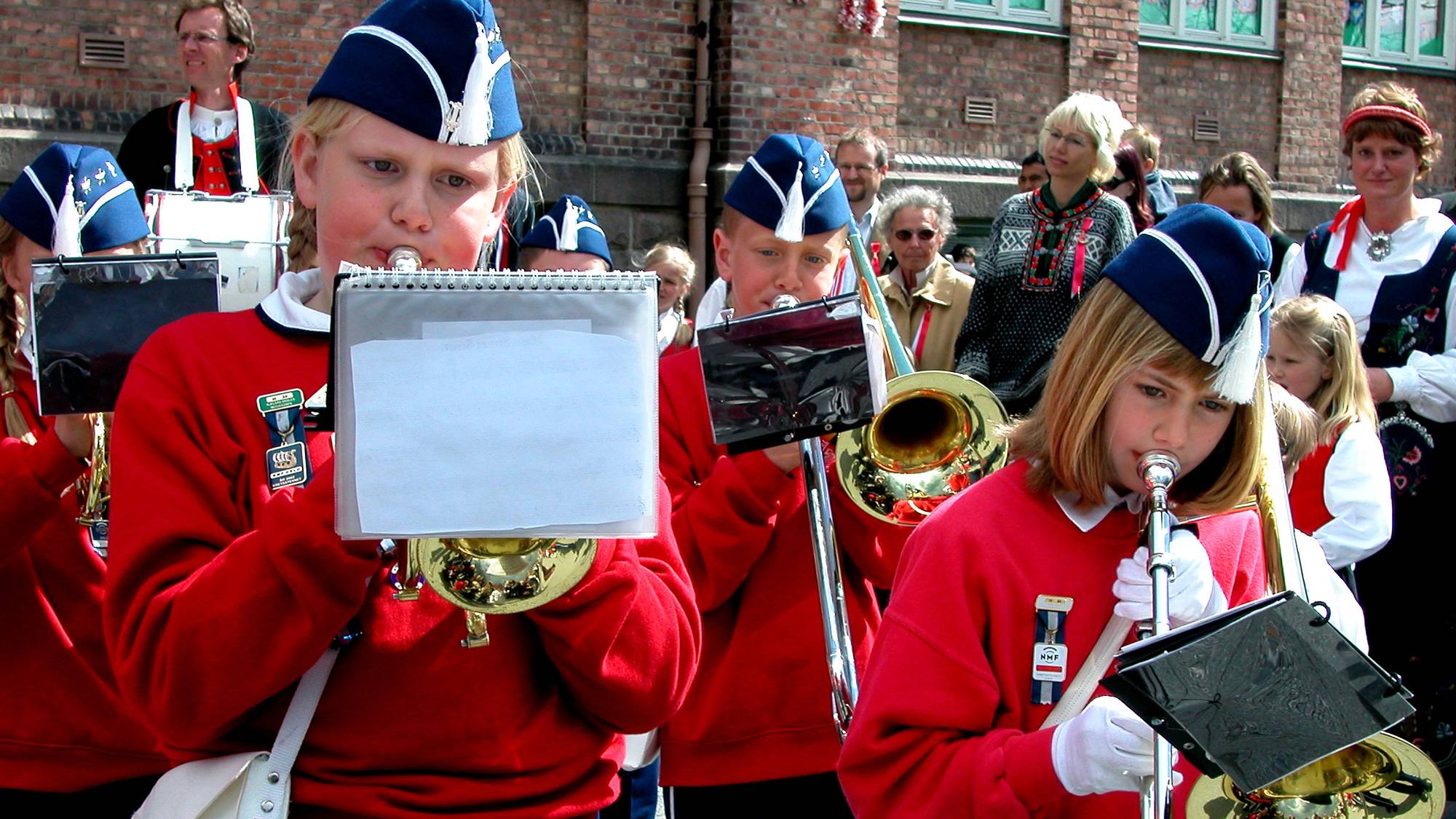 skolekorps. To jenter og en gutt i røde uniformer spiller kornett og tromboner, på 17.mai. Modellklarert.